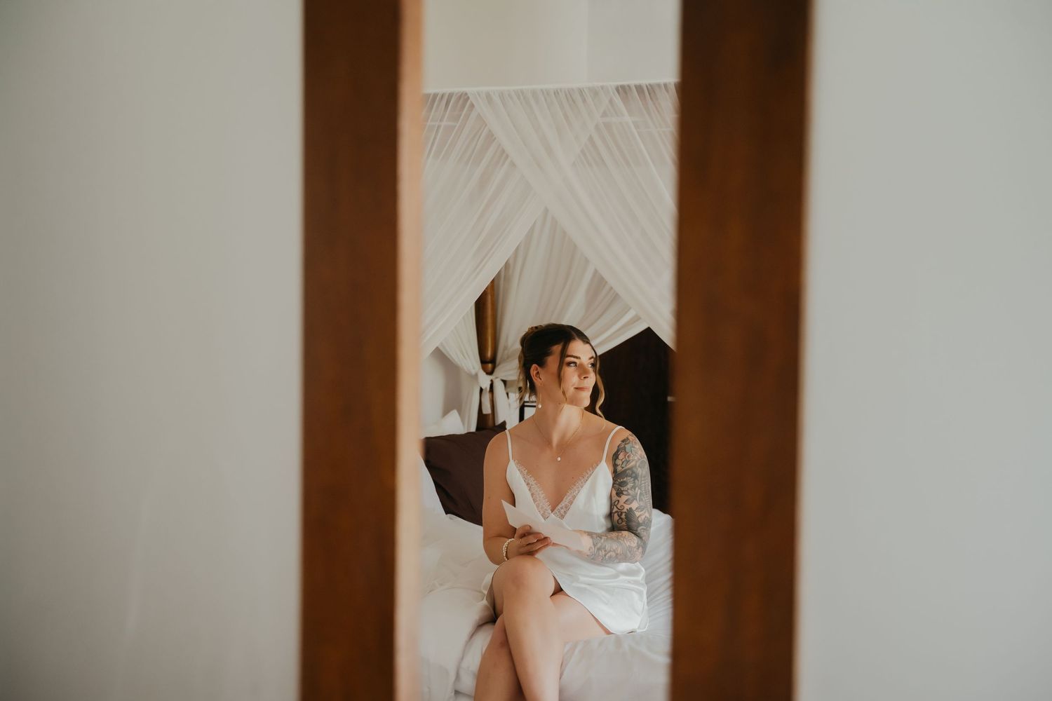 Bride in white dress sitting on bed viewed through mirror reflection.