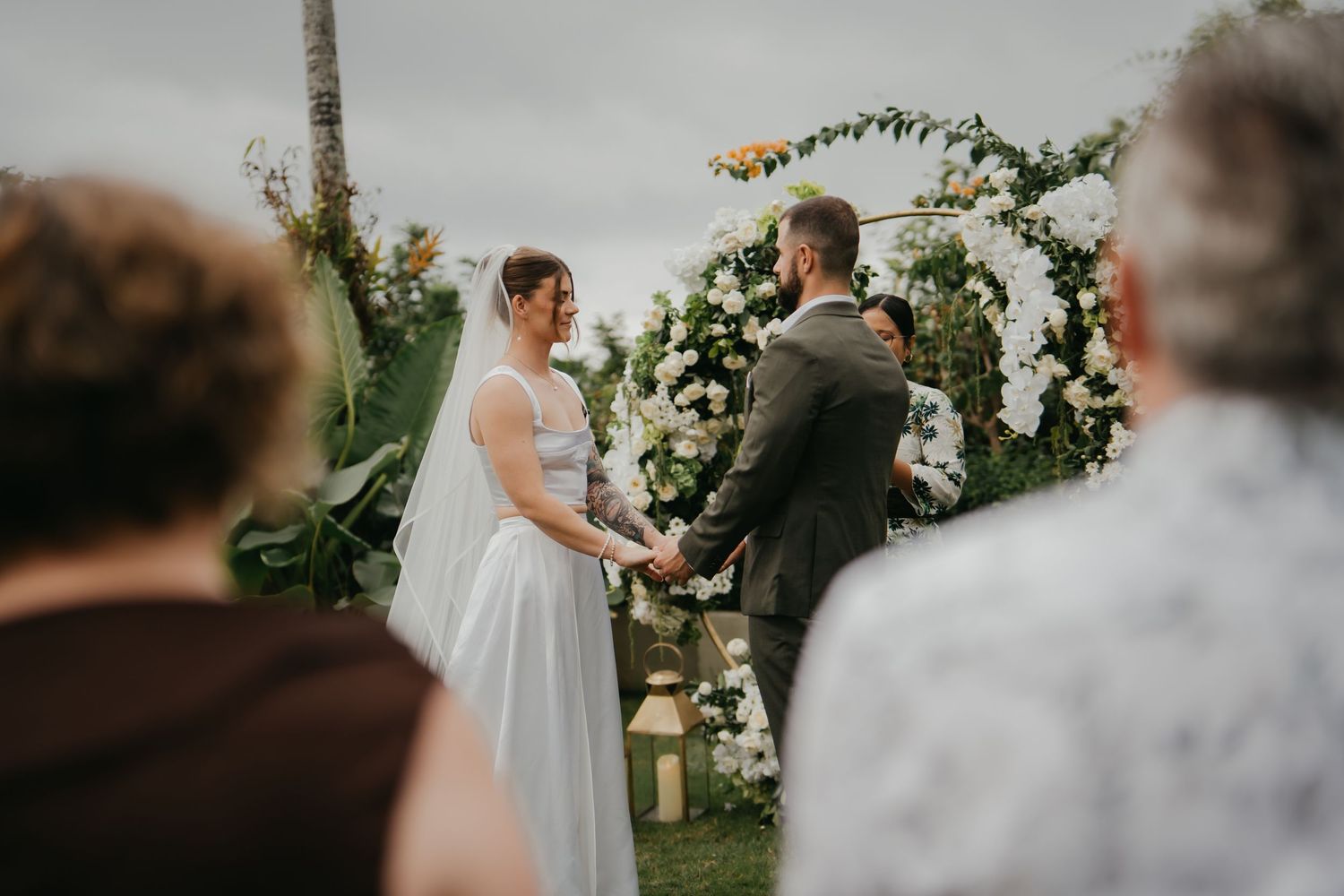 Couple holds hands at outdoor wedding altar surrounded by white flowers and guests.