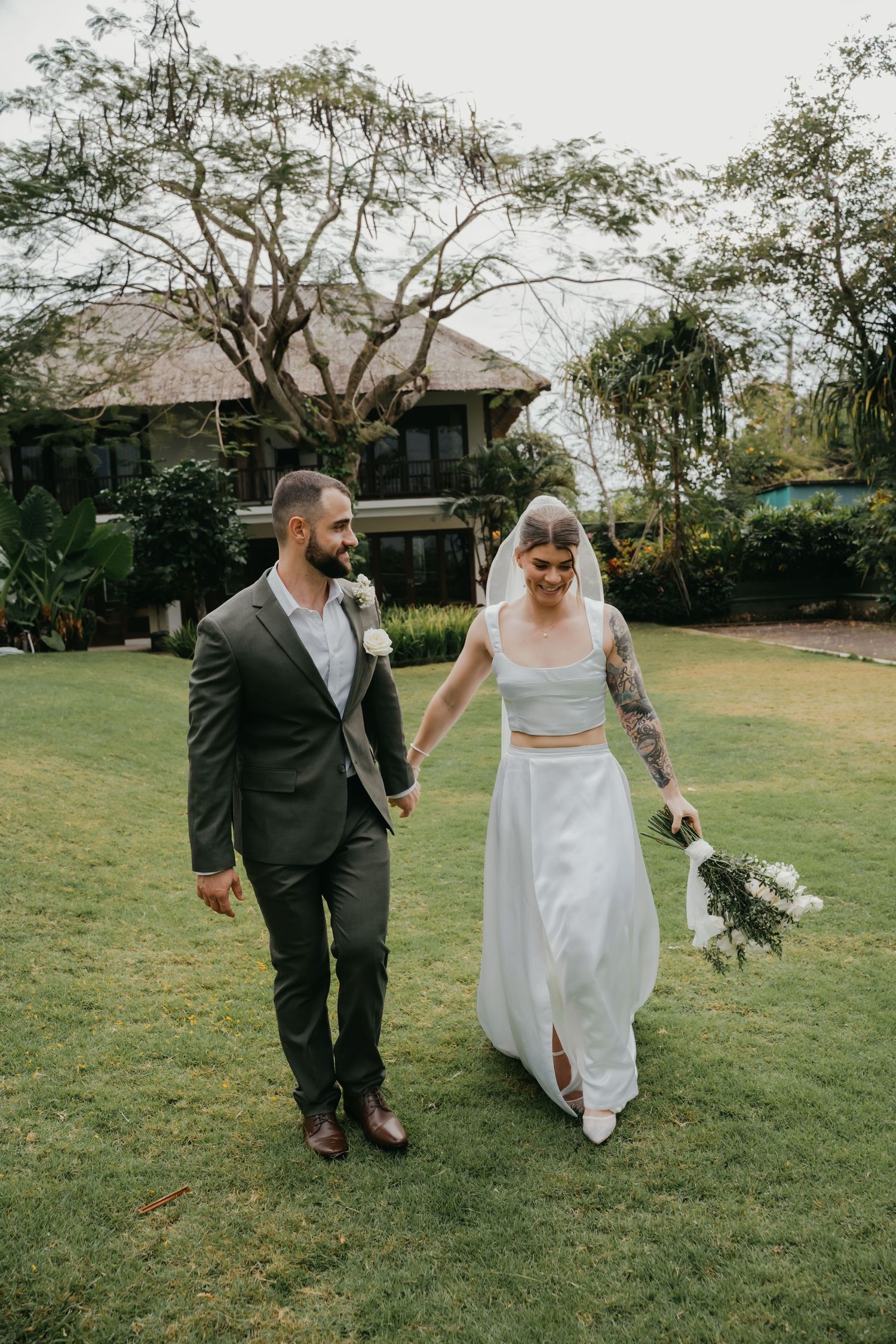 Bride and groom walk hand in hand across lush green lawn at tropical wedding venue.