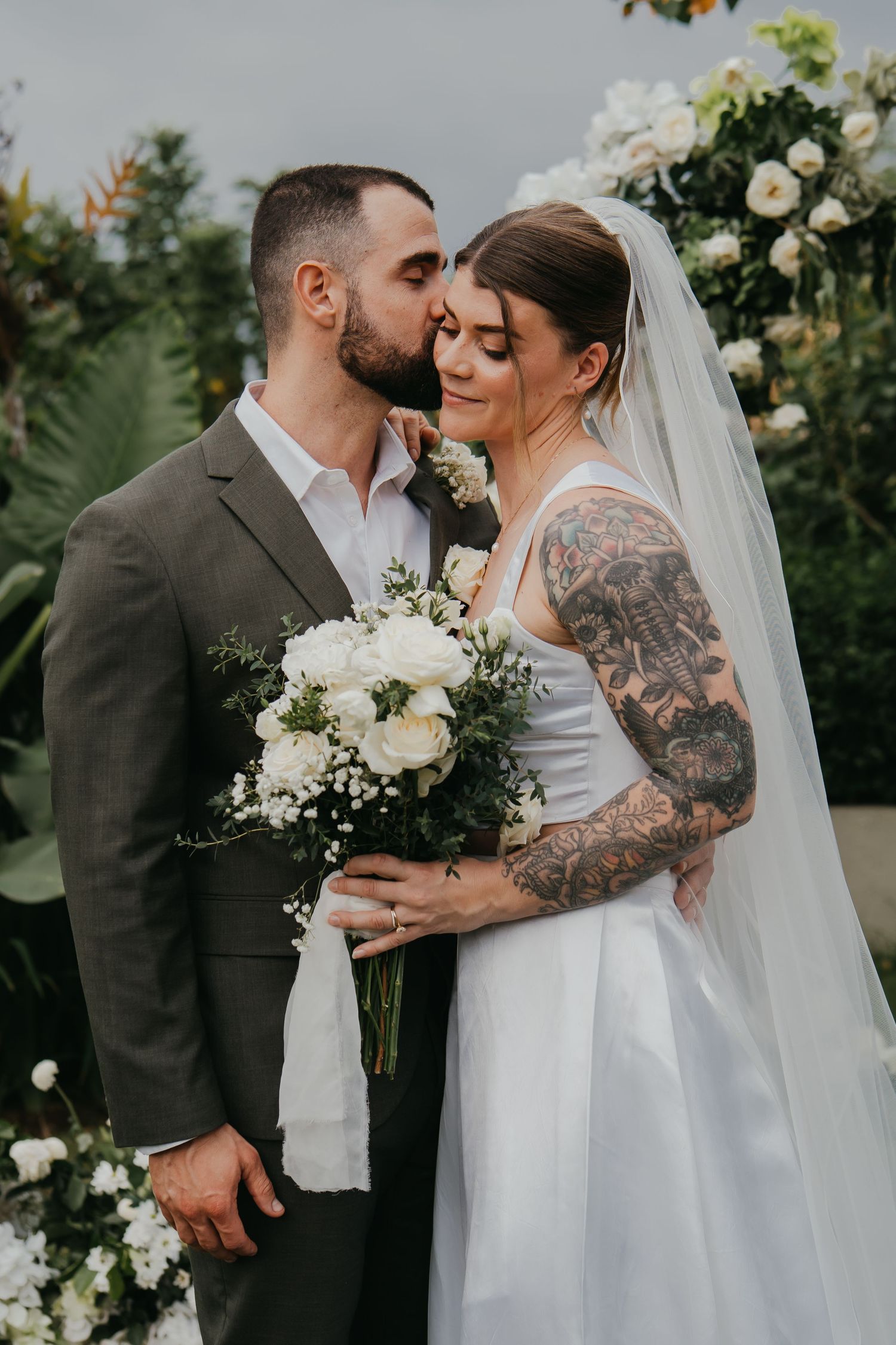 Groom kisses tattooed bride on forehead as she holds white rose bouquet before floral wedding arch.