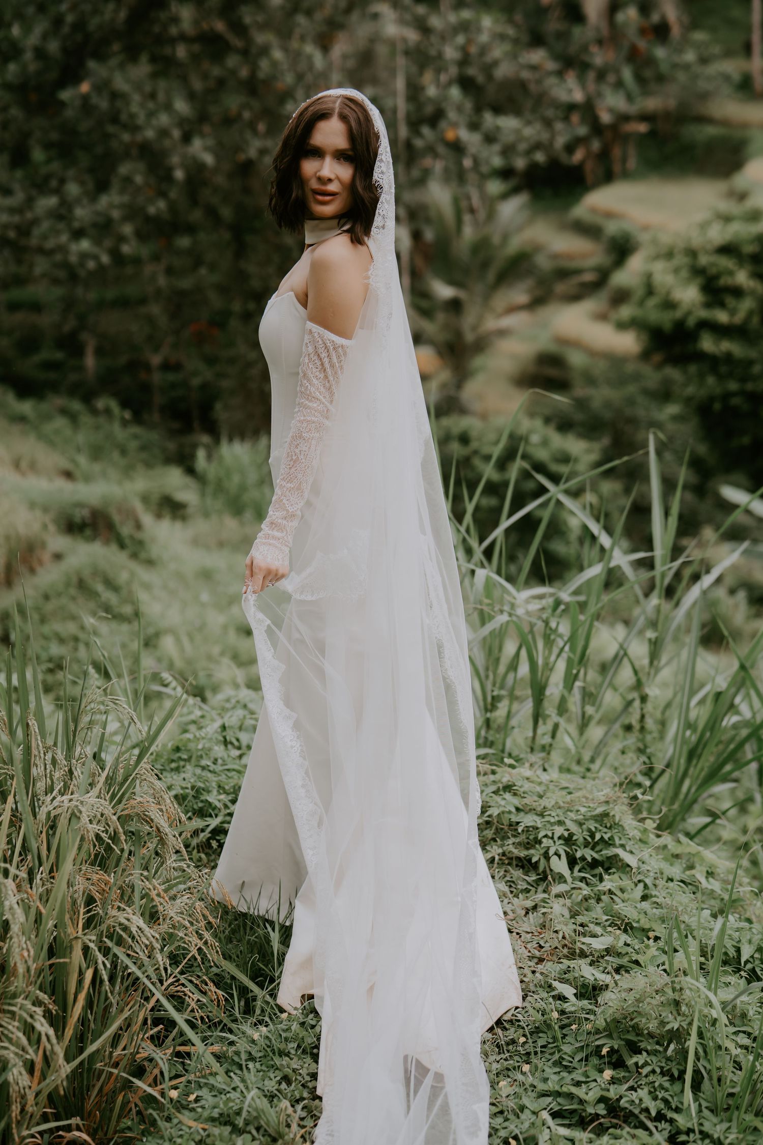 A bride in a flowing white wedding dress stands among lush green foliage in an outdoor garden setting.