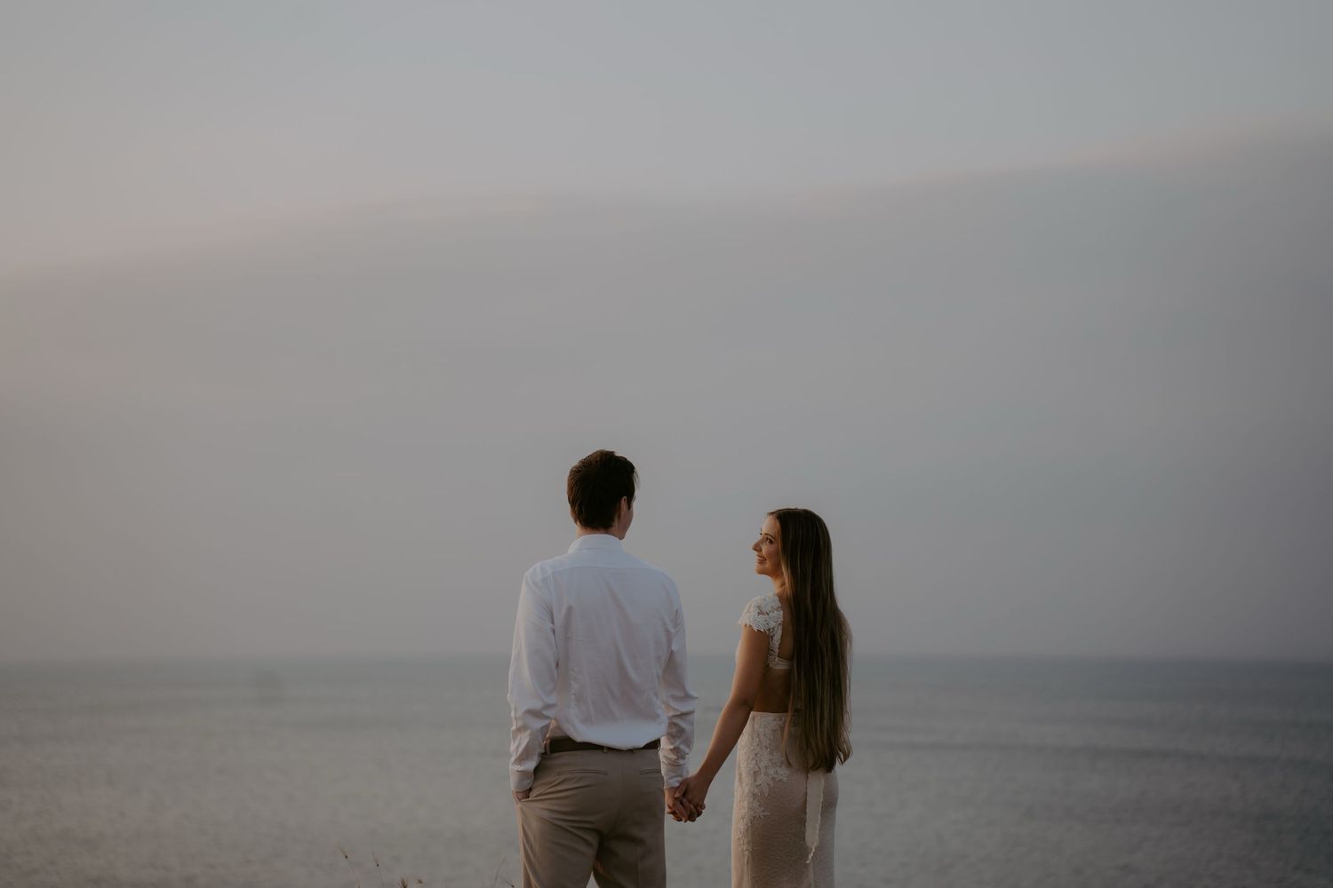 A couple in light colored clothing holds hands while looking out over a misty ocean view at dusk.