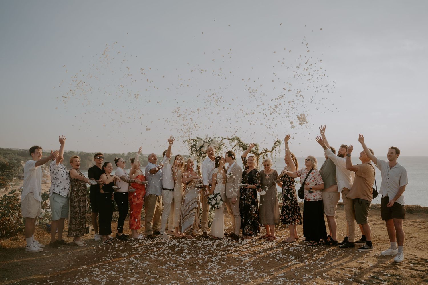 A group of wedding guests raise their arms in celebration against a scenic outdoor backdrop during golden hour.