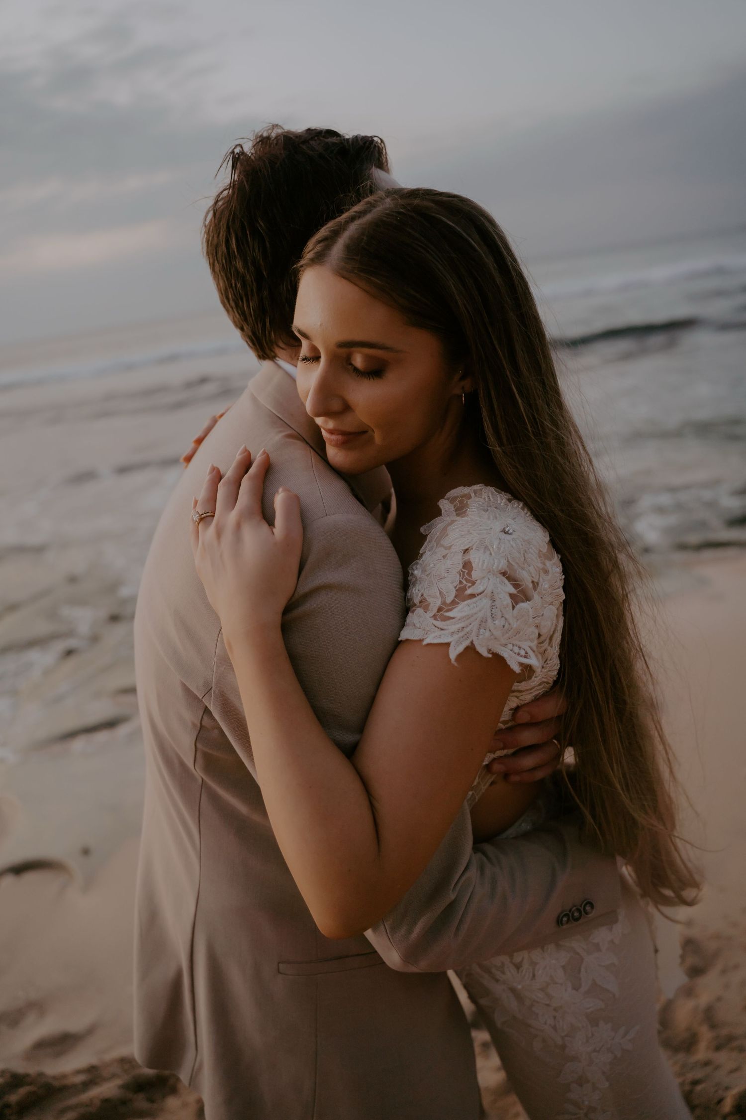 A romantic embrace between two people in wedding attire on a beach at sunset.