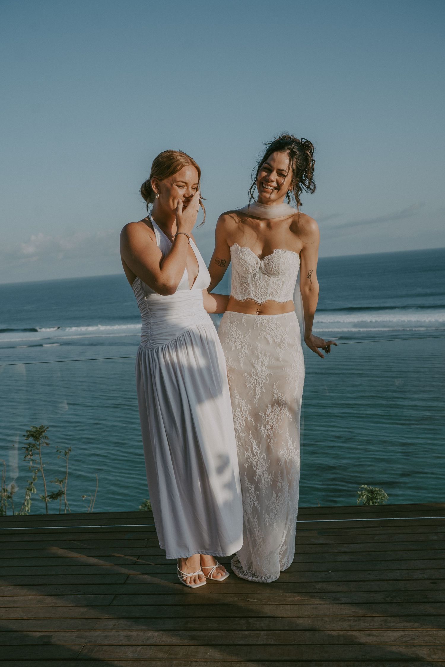 Two people in white dresses share a joyful moment on an oceanfront deck with blue waters and sky in the background.