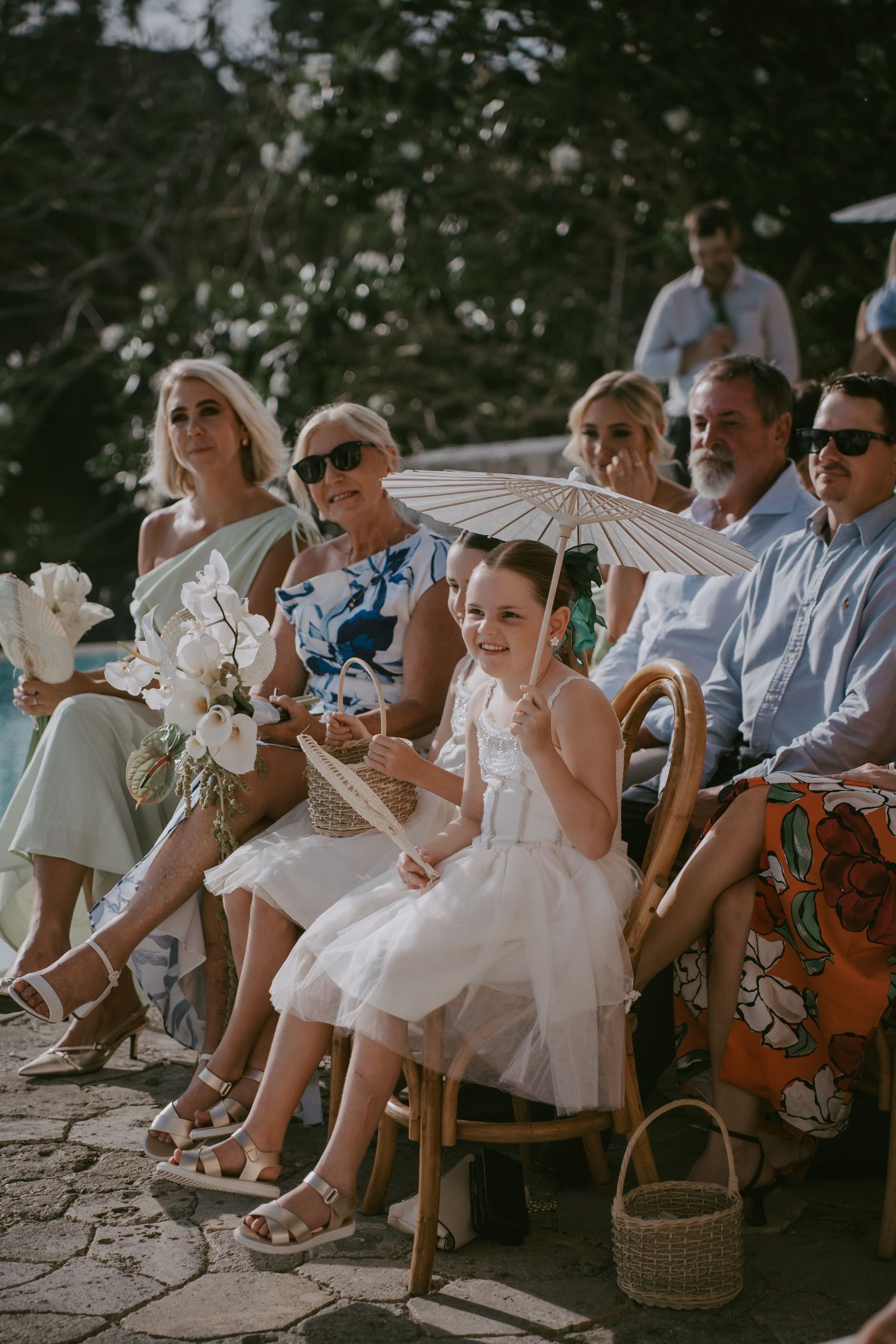 Wedding guests seated on rustic chairs holding white parasols during an outdoor ceremony.