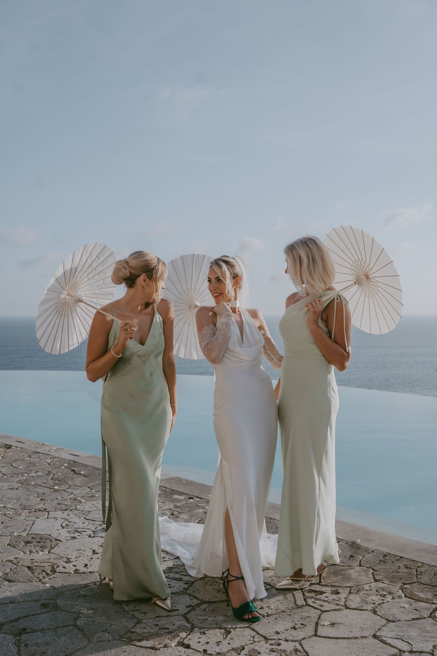 Three bridesmaids holding white parasols pose against an ocean backdrop at a cliffside wedding venue.
