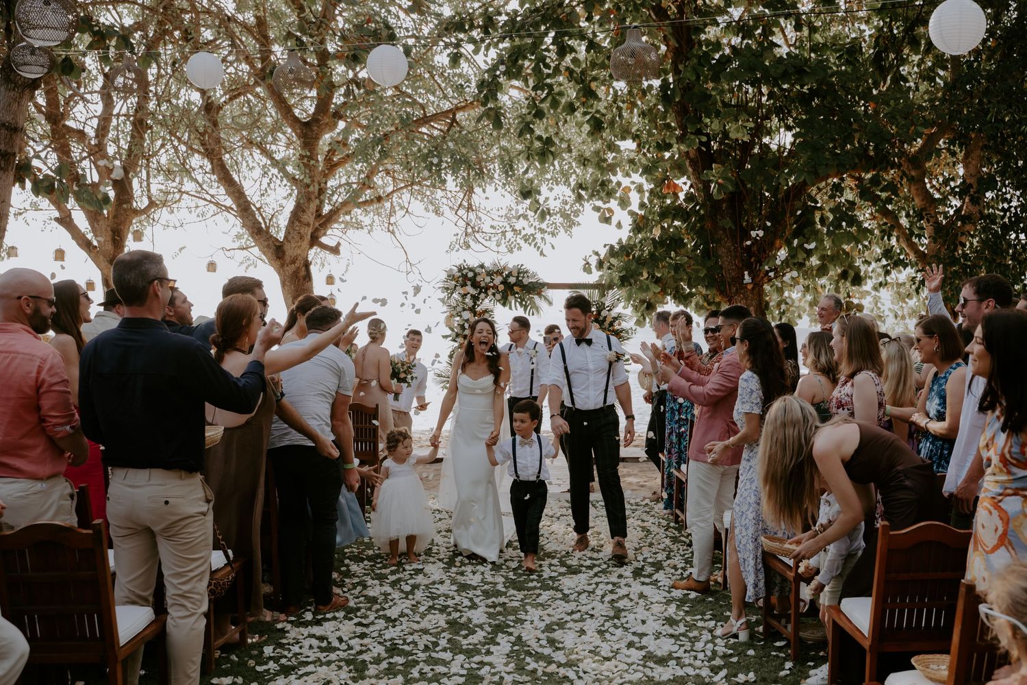 Wedding guests gather under trees during an outdoor ceremony as sunlight filters through the branches.