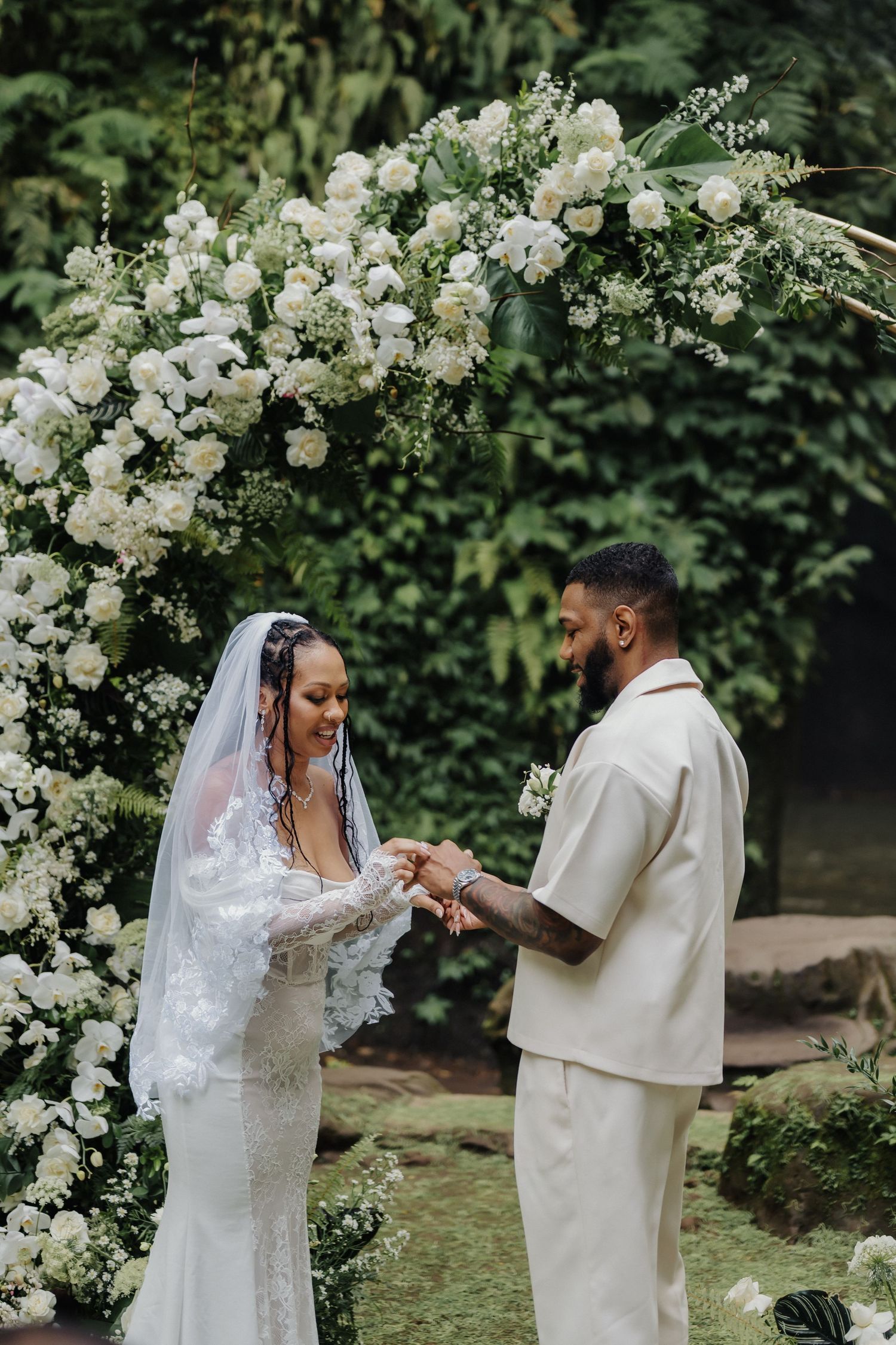 Bride and groom exchanging rings under a stunning white floral arch in a tropical garden wedding.