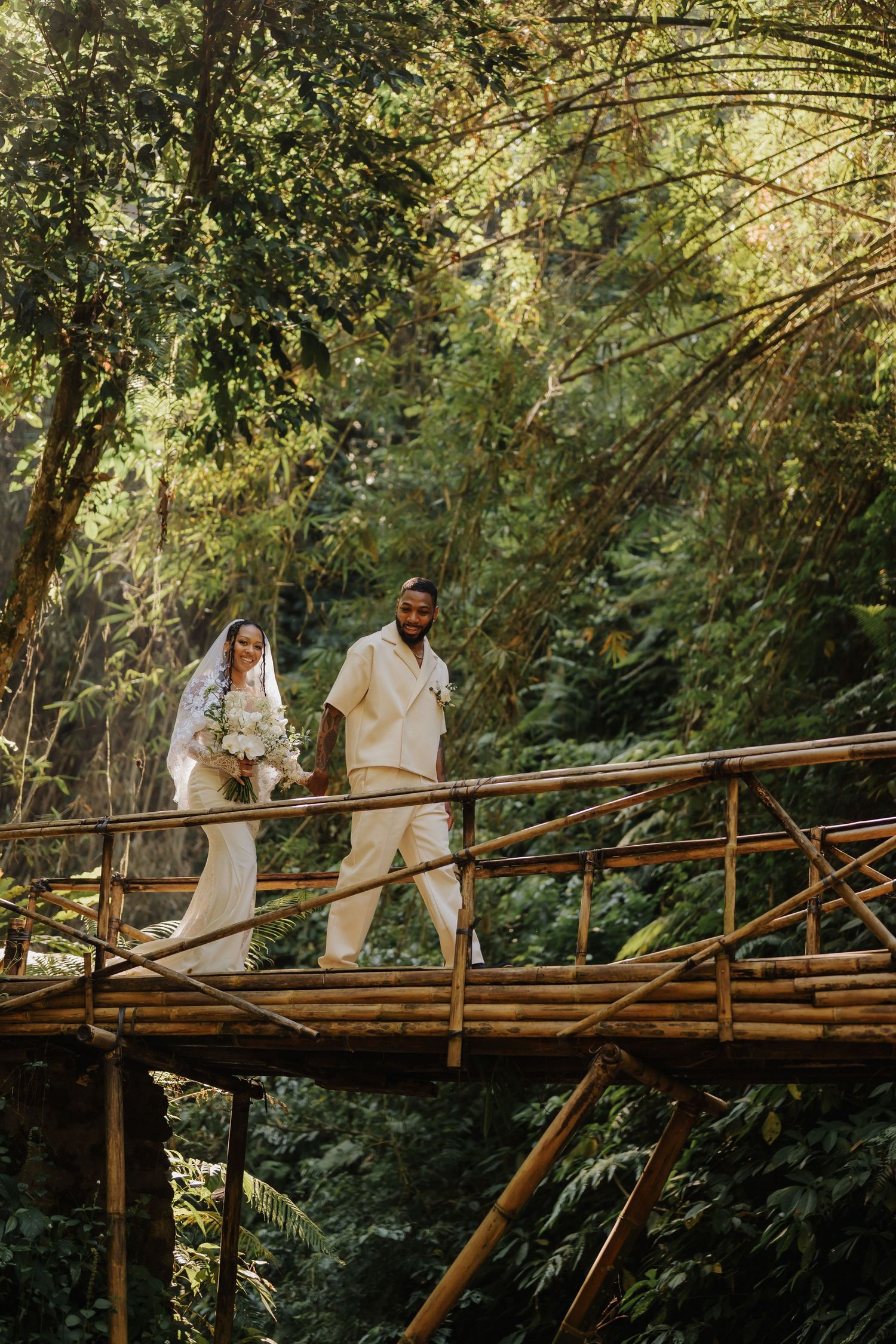 Bride and groom walk hand in hand across a bamboo bridge in a sunlit tropical forest.