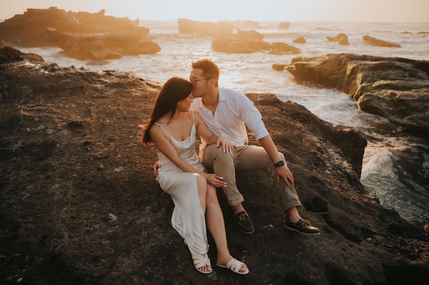 Couple sitting on rocky coastal cliffs at golden sunset, sharing a romantic moment with ocean waves in the background.