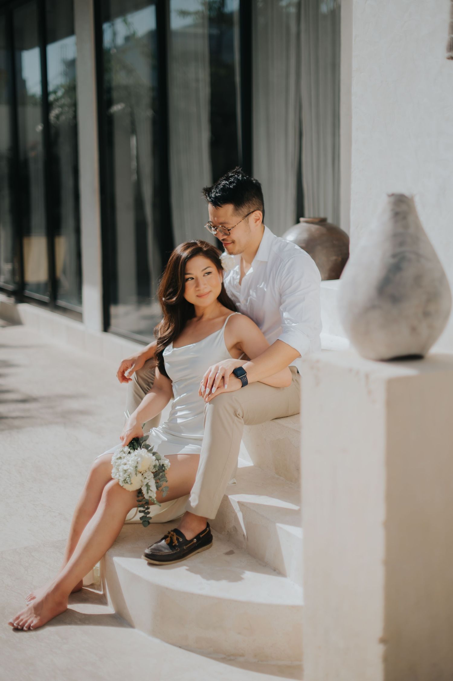 Couple in white outfits sitting on steps with a bouquet, sharing a romantic moment outdoors.