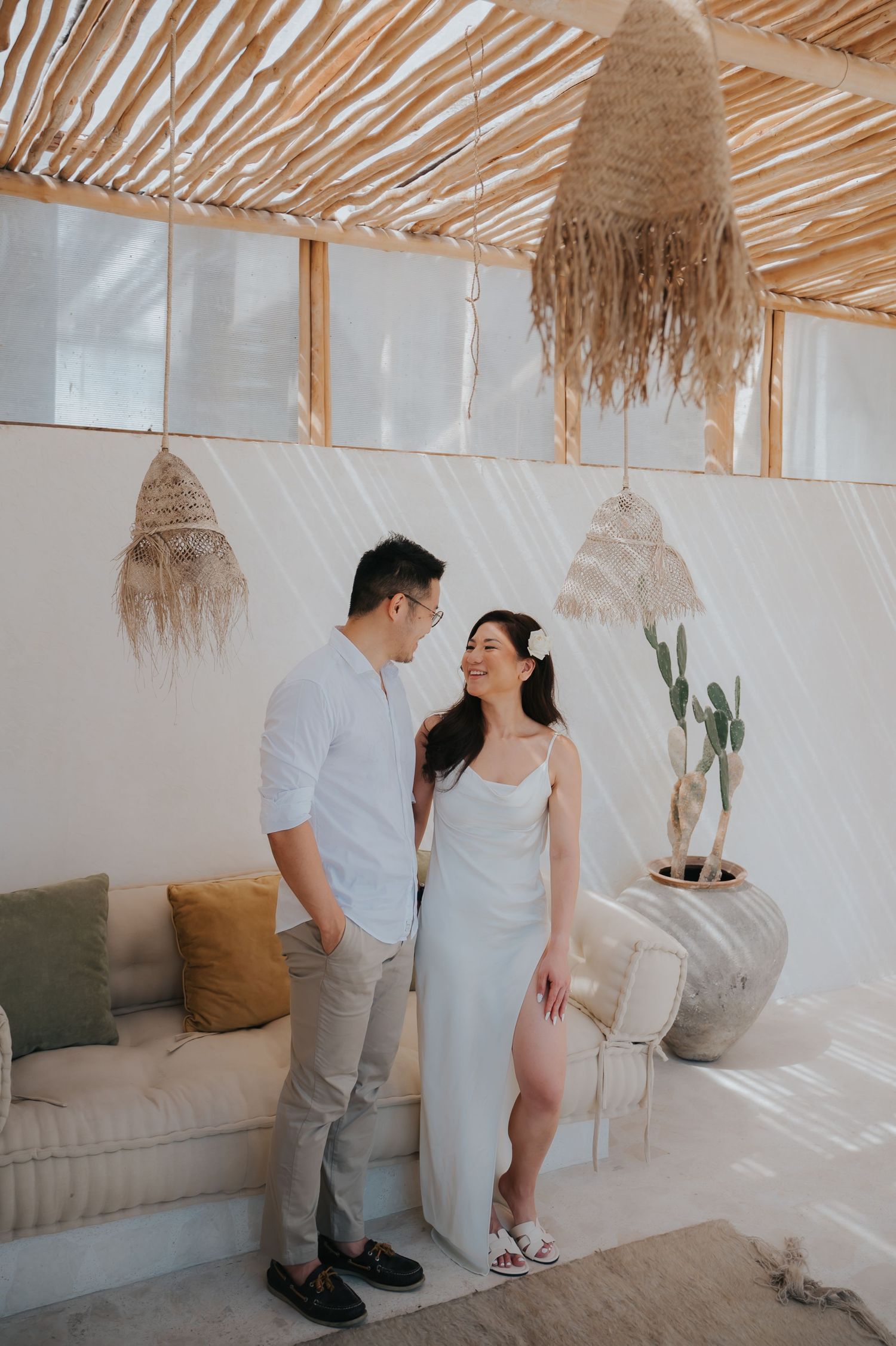 Couple in boho beach cabana, man in white shirt and woman in white slip dress, sharing a romantic moment.