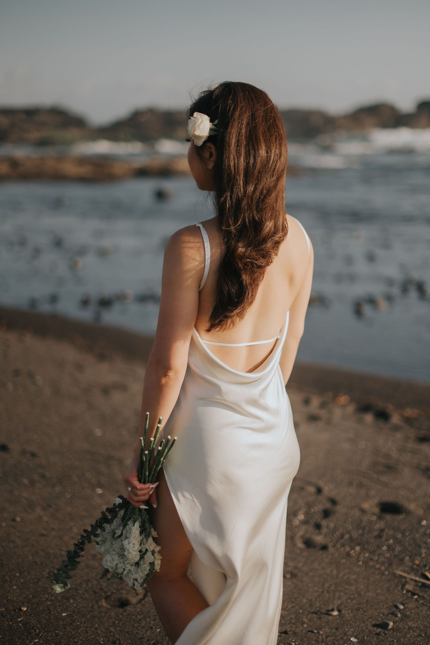 Bride in white backless satin gown holding bouquet on sandy beach at sunset with ocean waves in background.