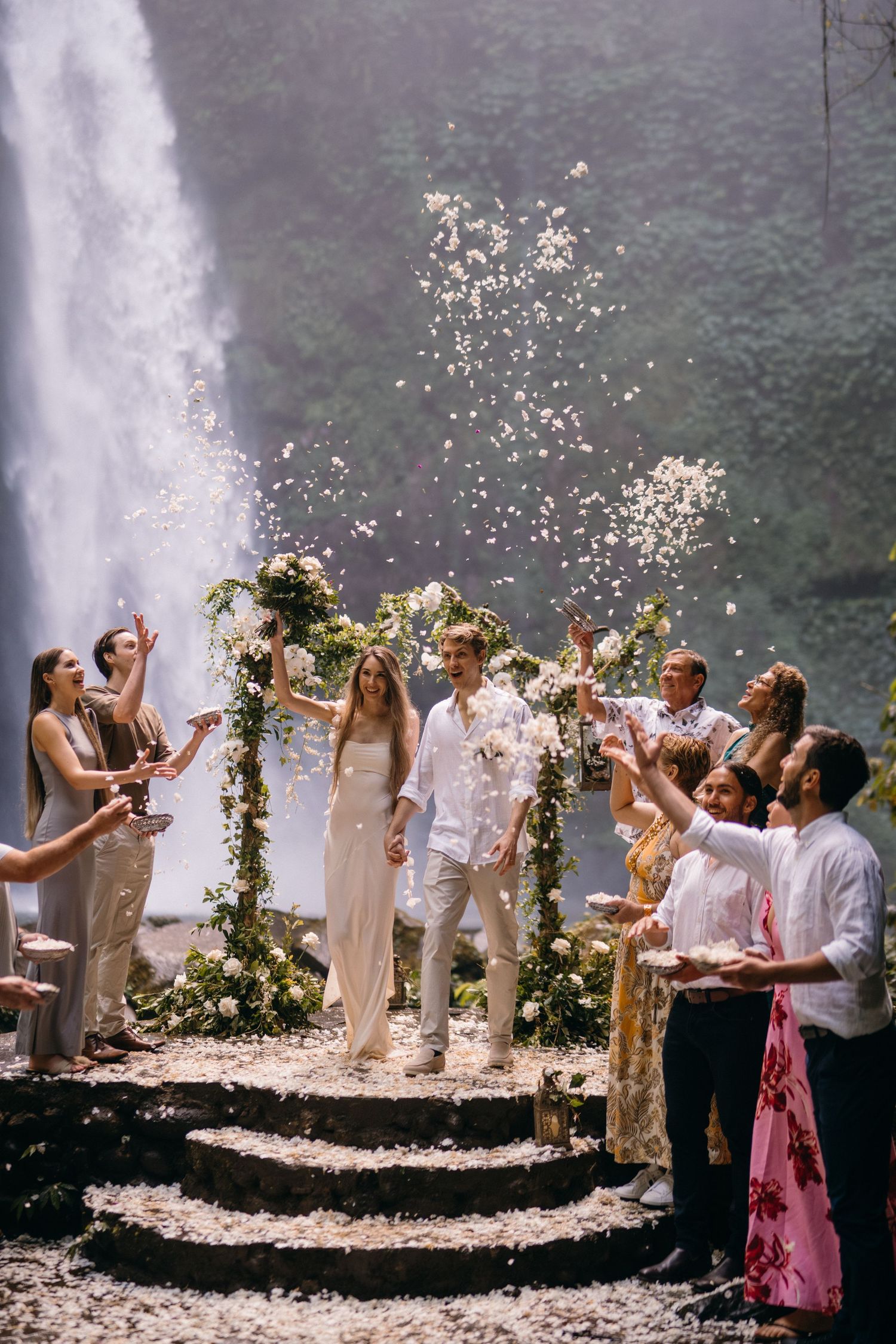 A wedding celebration near a waterfall with people throwing confetti while standing on stone steps adorned with floral decorations.