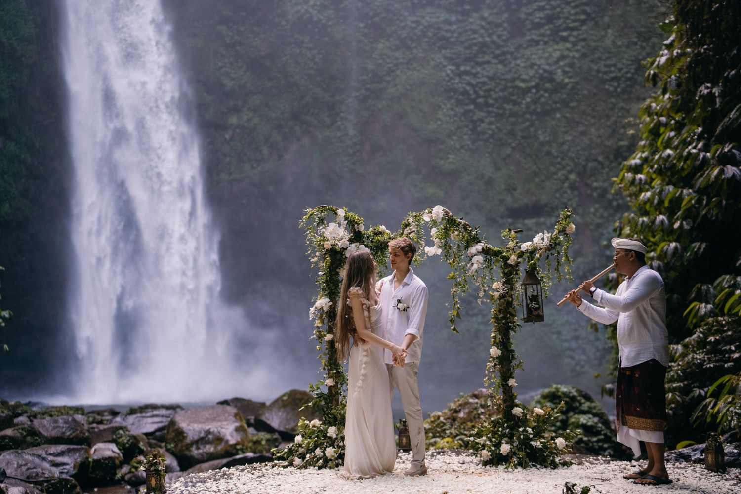 A couple poses in front of a majestic waterfall in a lush tropical setting during their wedding ceremony.