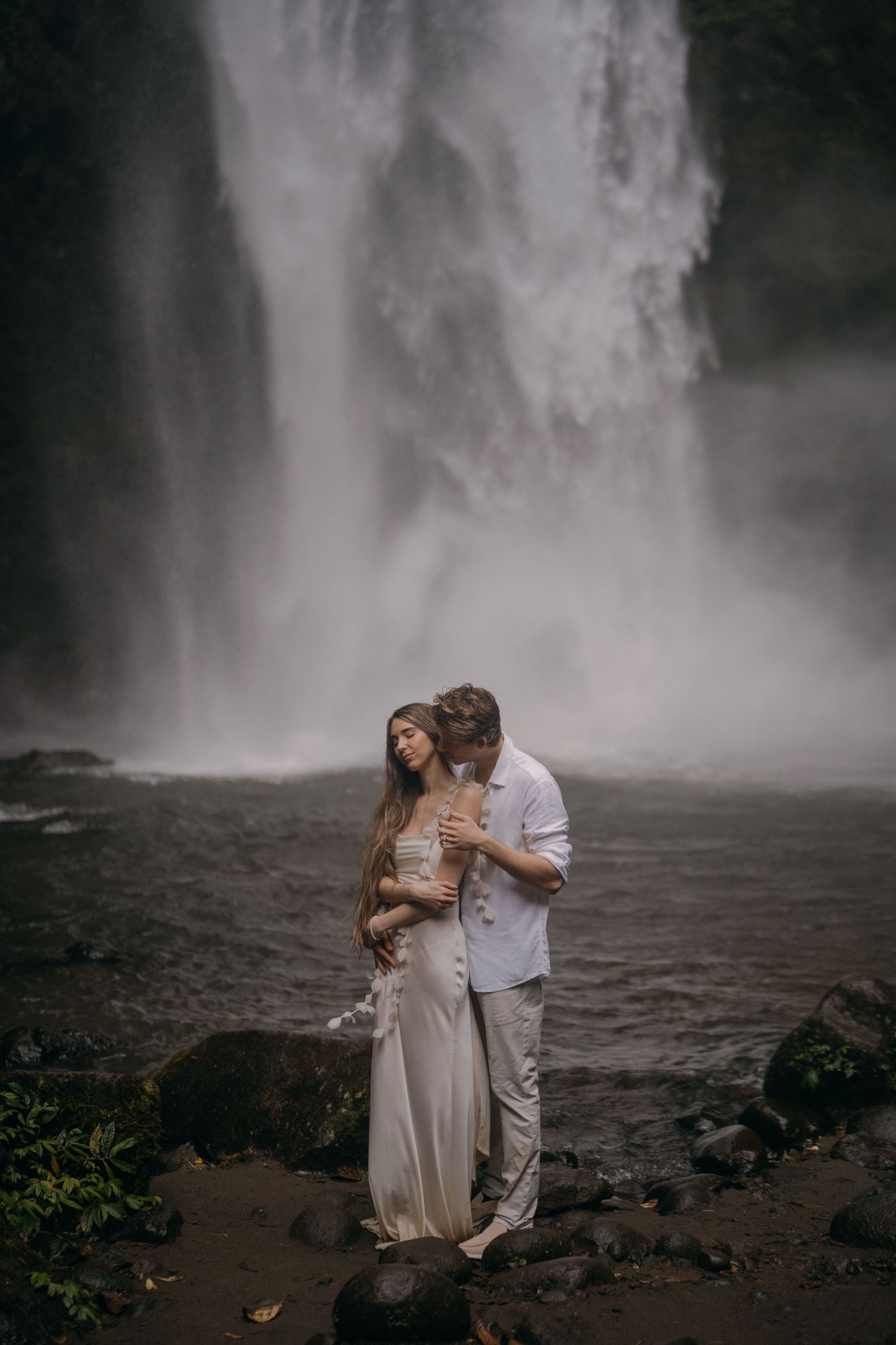 A romantic couple embraces near a misty waterfall with dark rocks in the foreground, creating a dramatic natural backdrop.