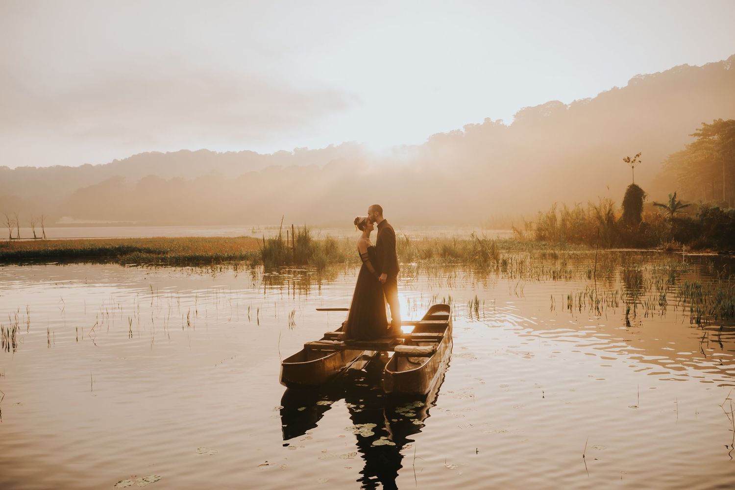 Silhouetted boats float peacefully on a misty lake during golden hour with mountains in the background.
