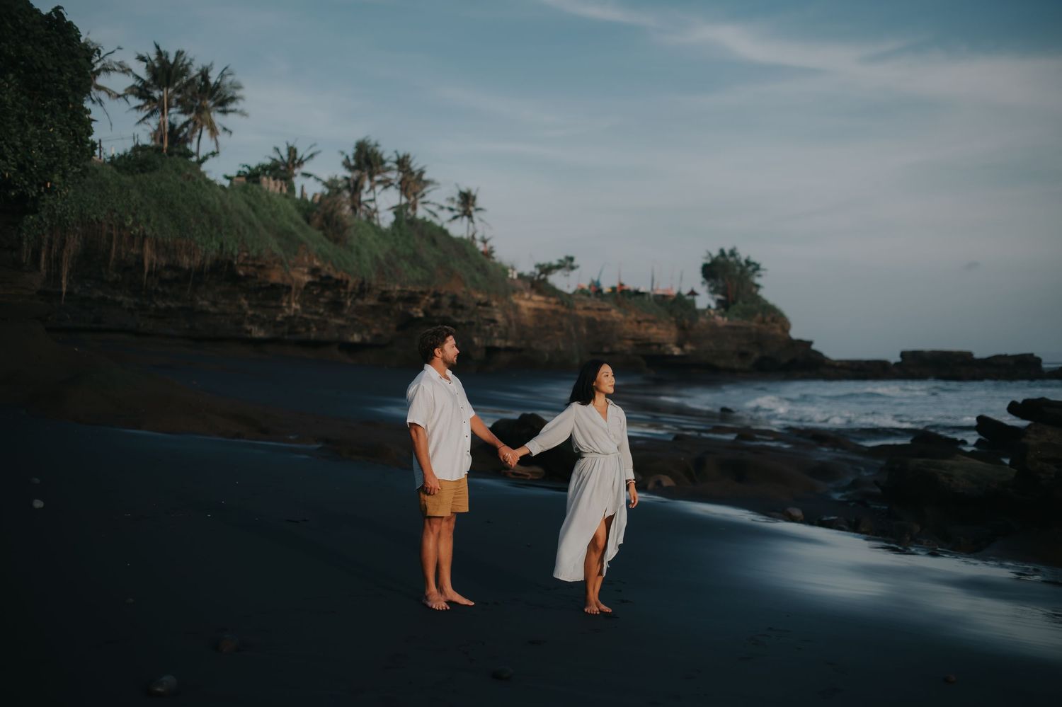 Silhouetted figures walk along a scenic black sand beach at dusk with palm trees and cliffs in the background.