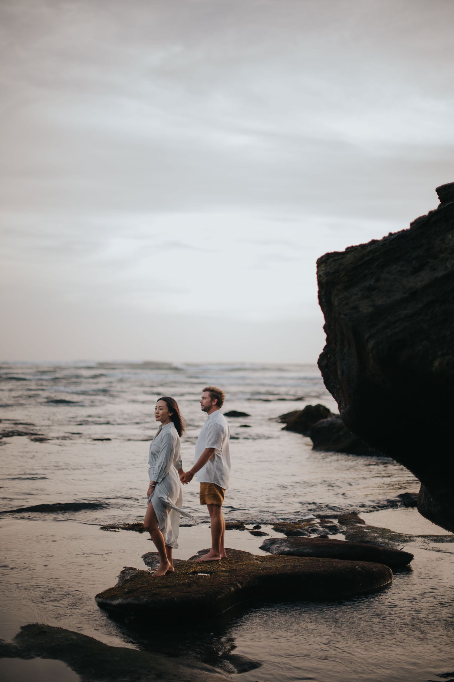 A couple sits together on a rocky outcrop overlooking a moody beach at sunset.