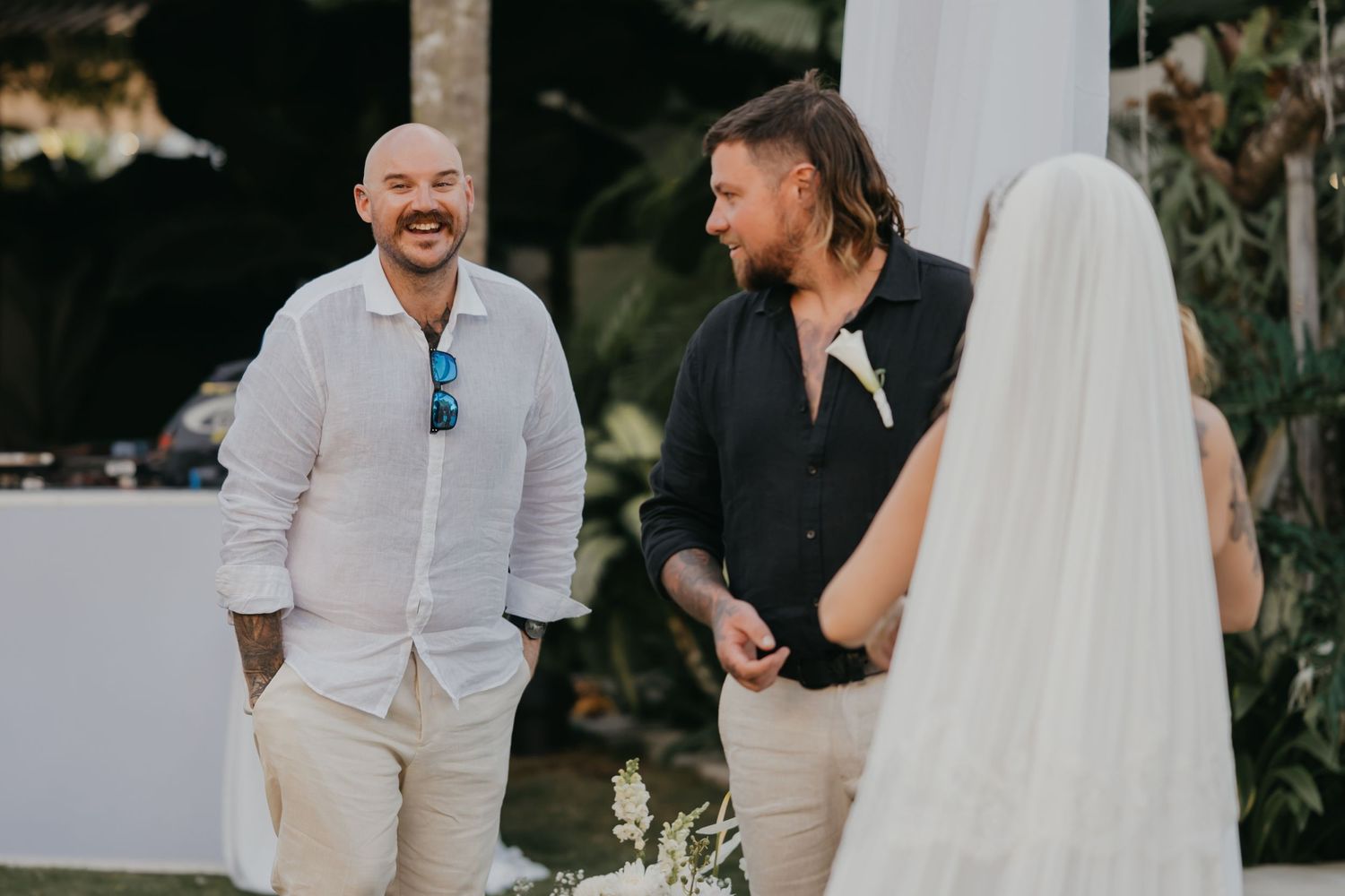 Groom in black shirt laughing with guest in white shirt while bride in veil approaches tropical wedding ceremony.