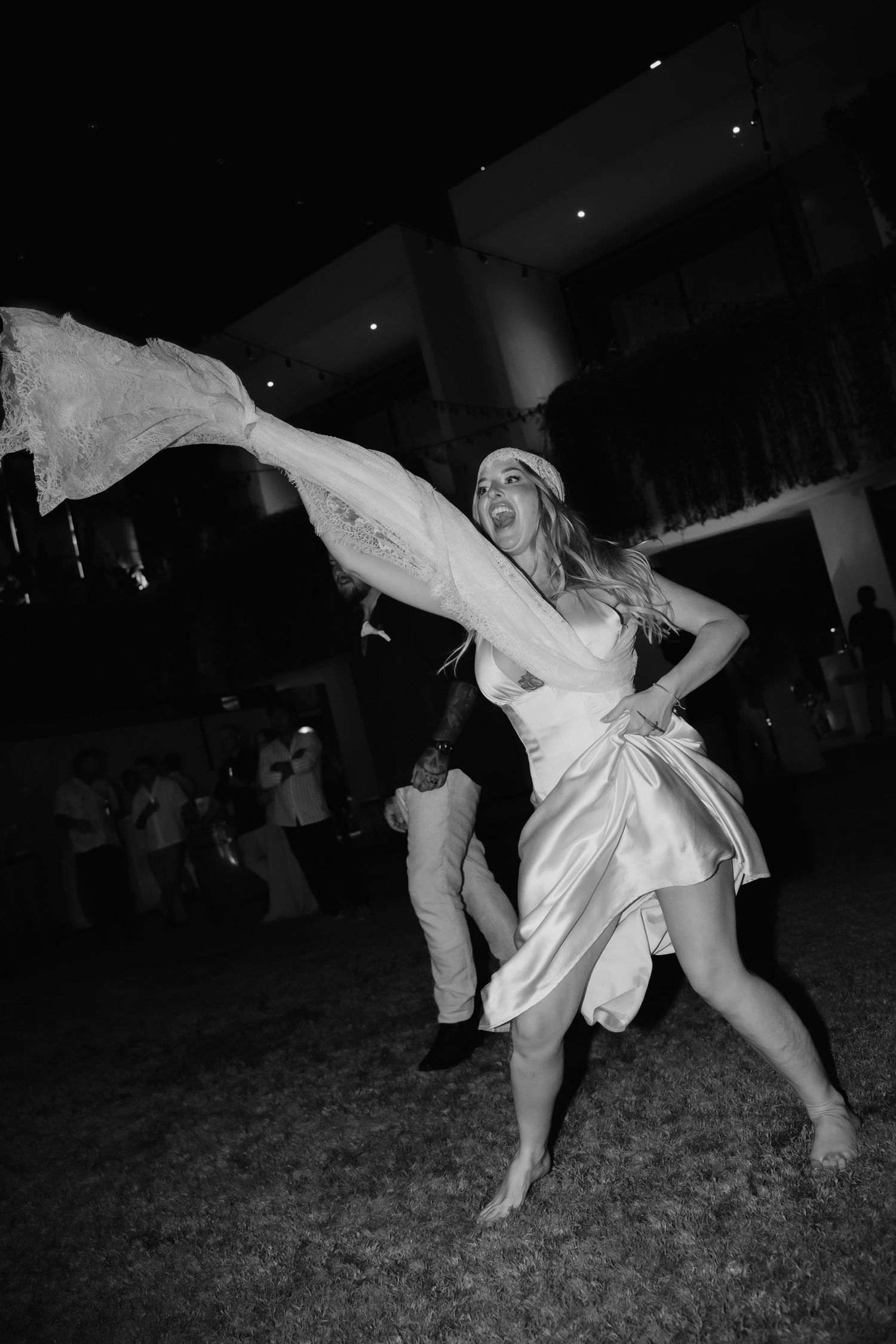 Black and white photo of barefoot bride dancing energetically outdoors at her wedding reception.