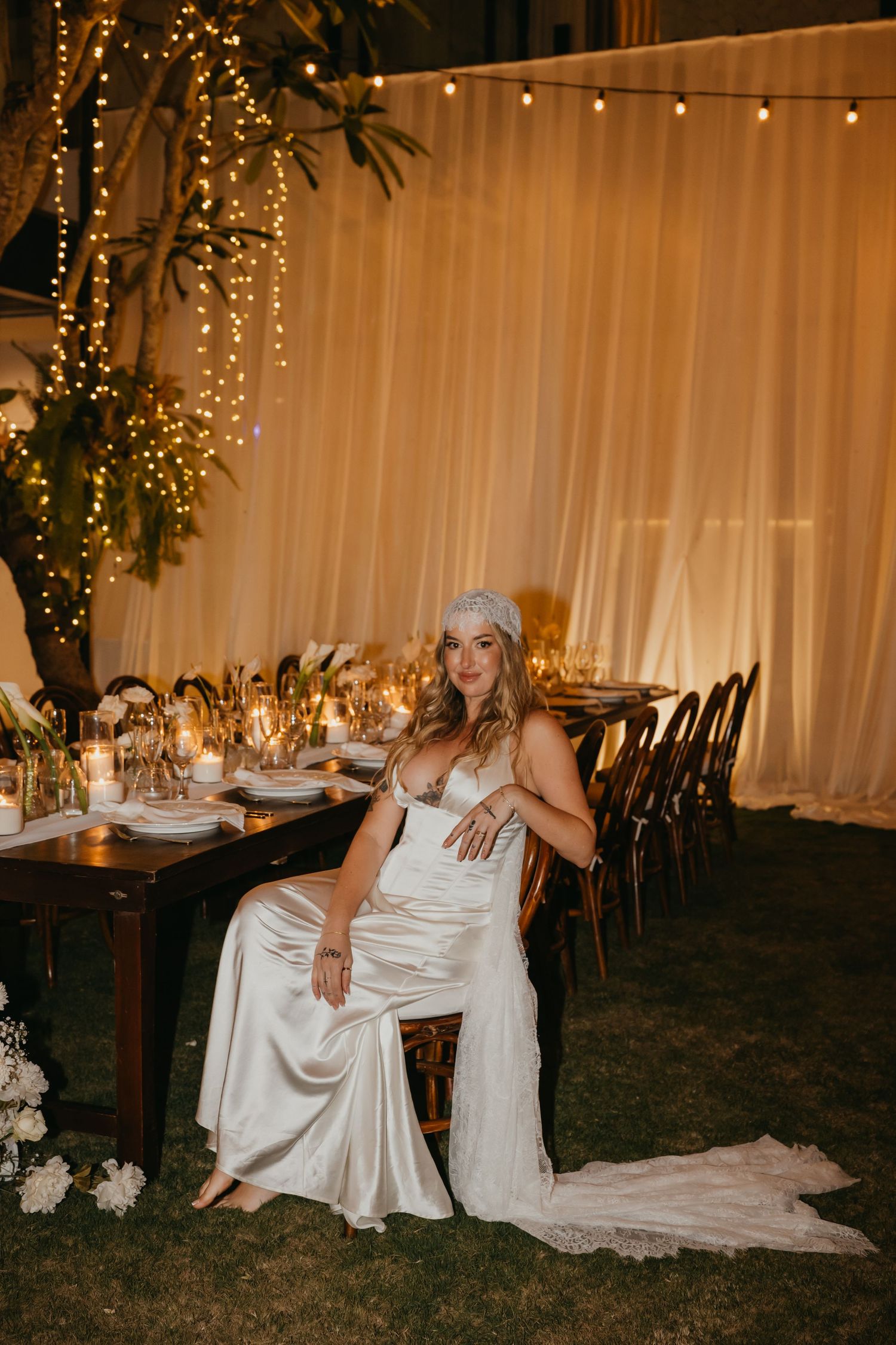 Bride in satin gown sitting at decorated wedding reception table surrounded by candlelight and fairy lights.