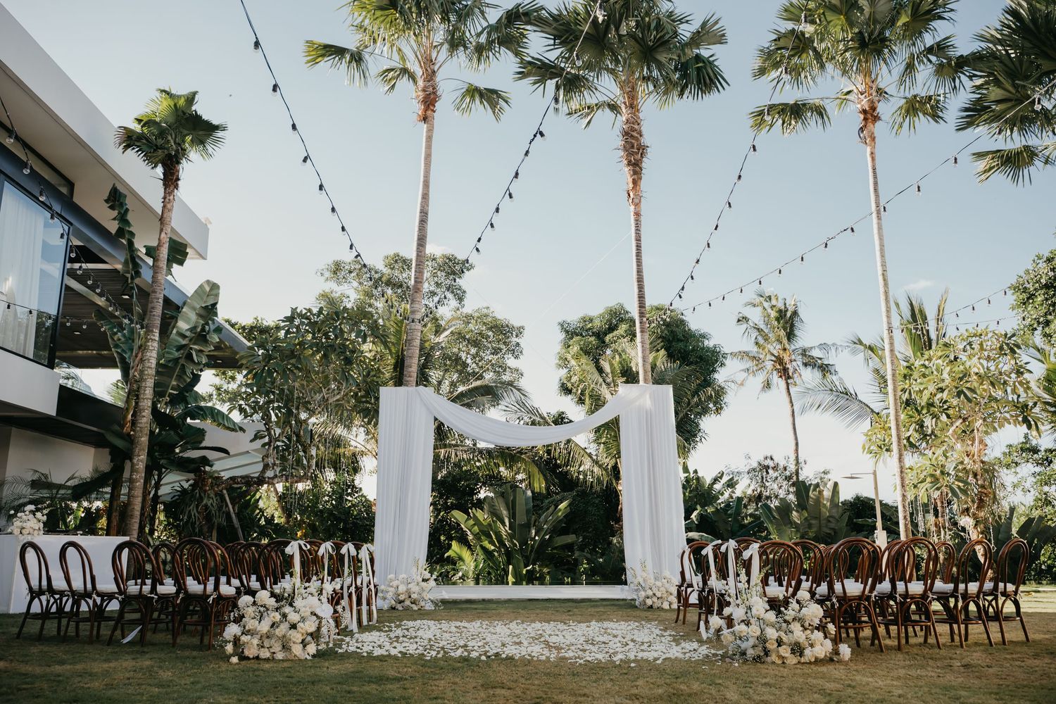 Elegant outdoor wedding ceremony setup with white arch, palm trees, floral aisle and string lights.
