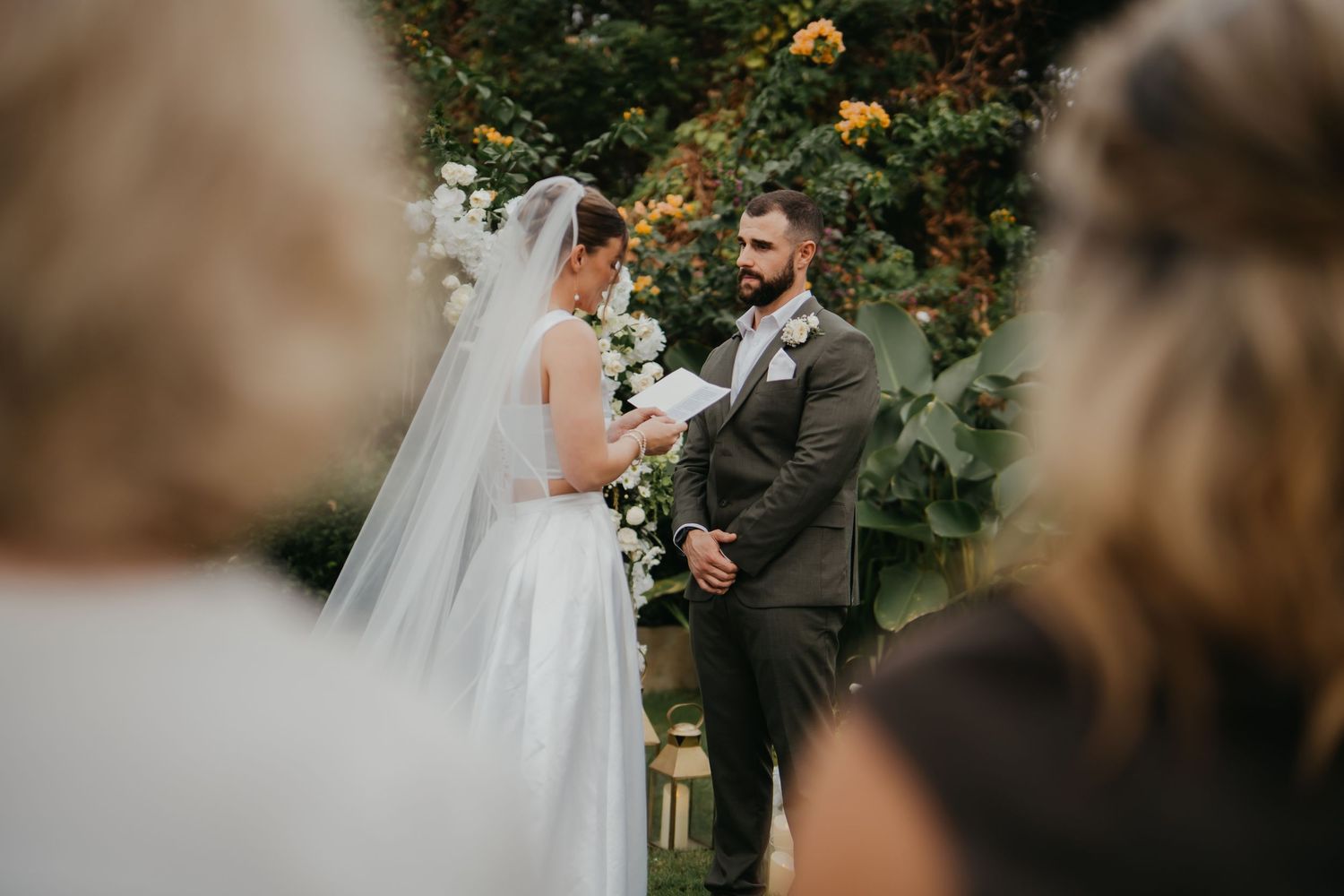 Groom gazing lovingly at veiled bride during outdoor wedding ceremony with white floral arch backdrop.