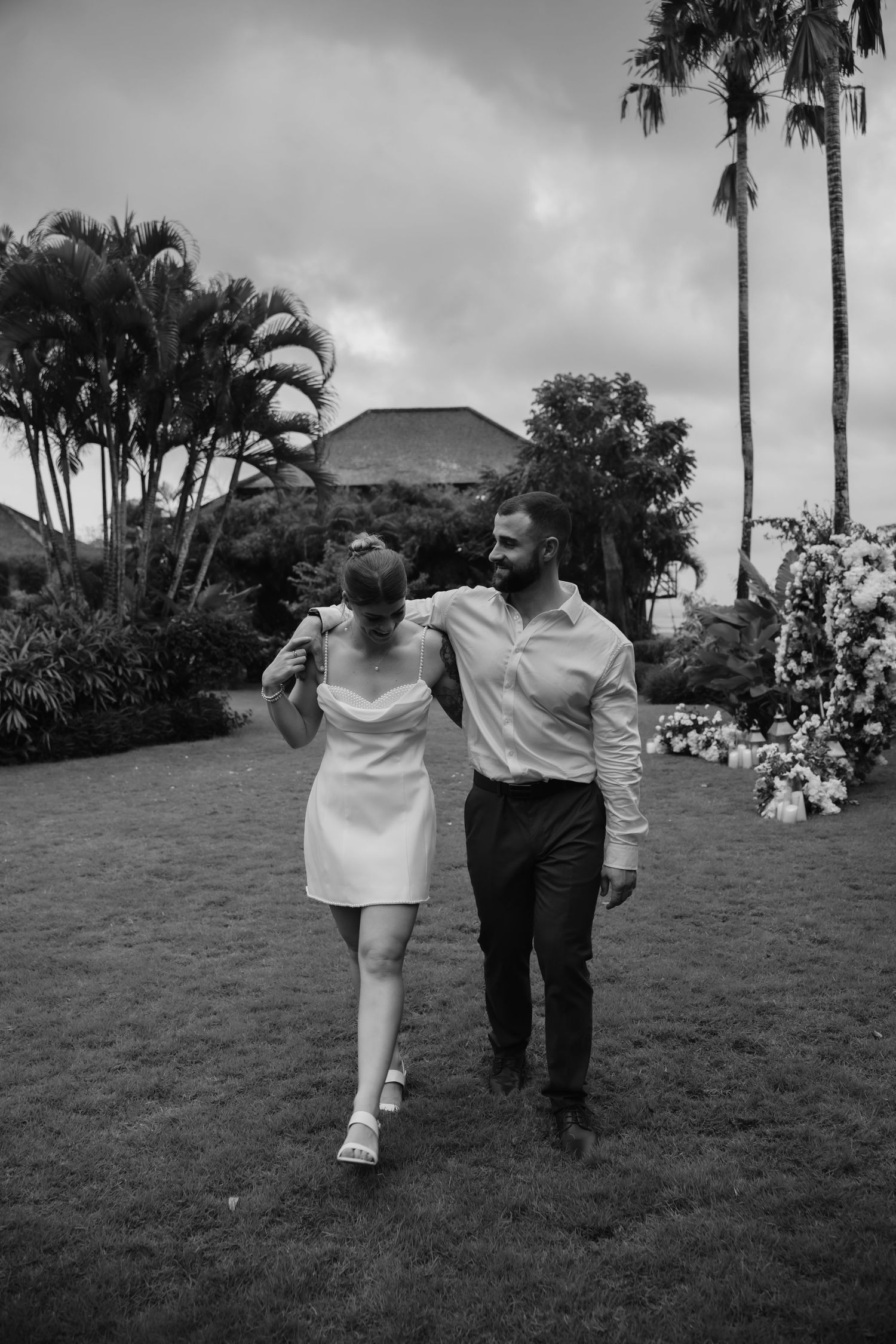 Black and white photo of smiling newlyweds walking arm in arm across tropical garden lawn.