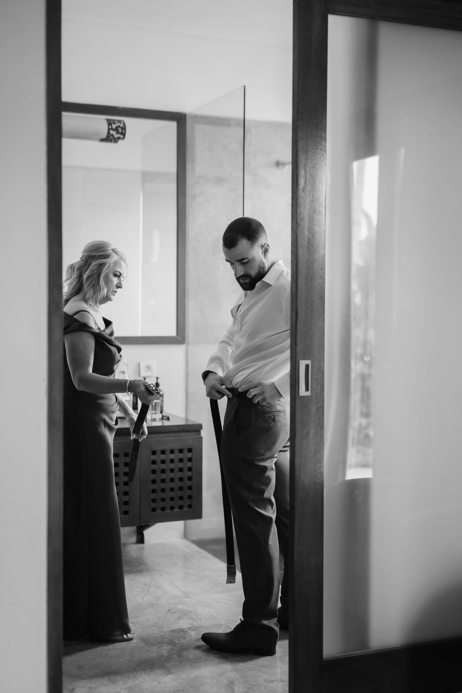 Black and white photo of a couple getting ready for a wedding, man adjusting his belt while woman assists.