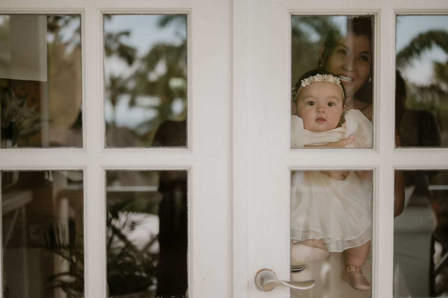 Adorable baby in white dress with floral headband peering through a white glass-paned door at a wedding.