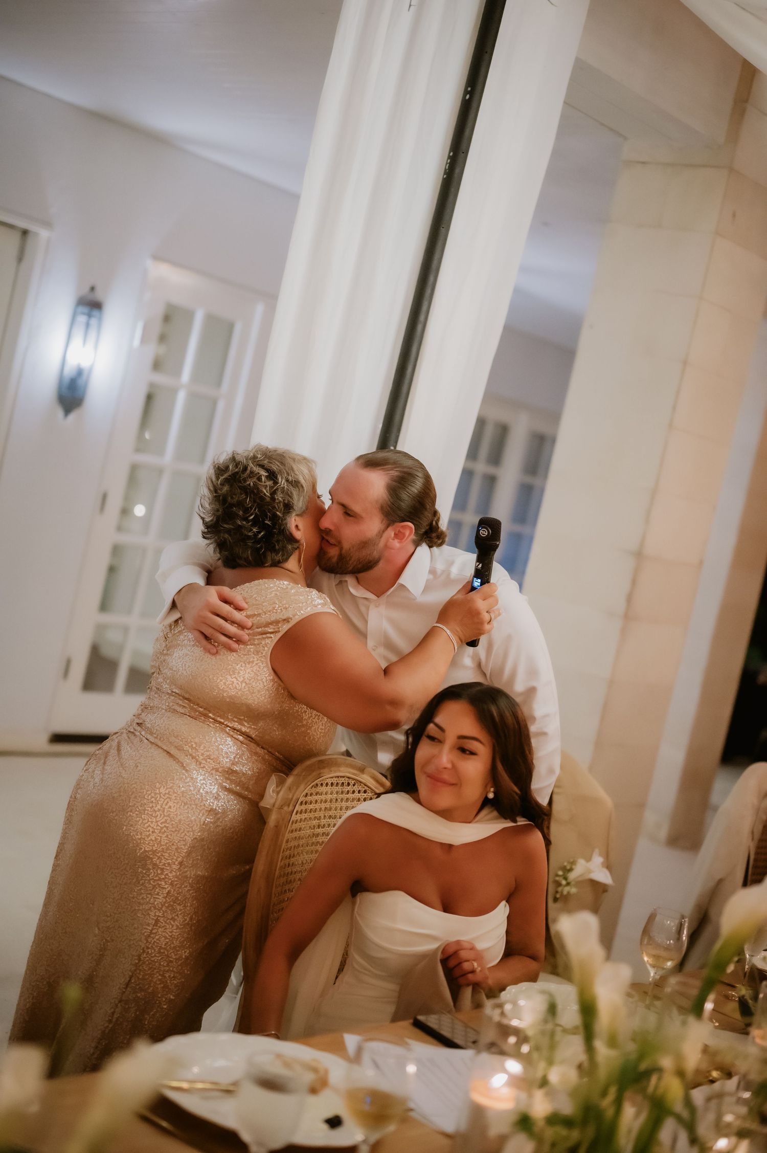 Groom kisses older woman while bride smiles at candlelit wedding reception dinner table.