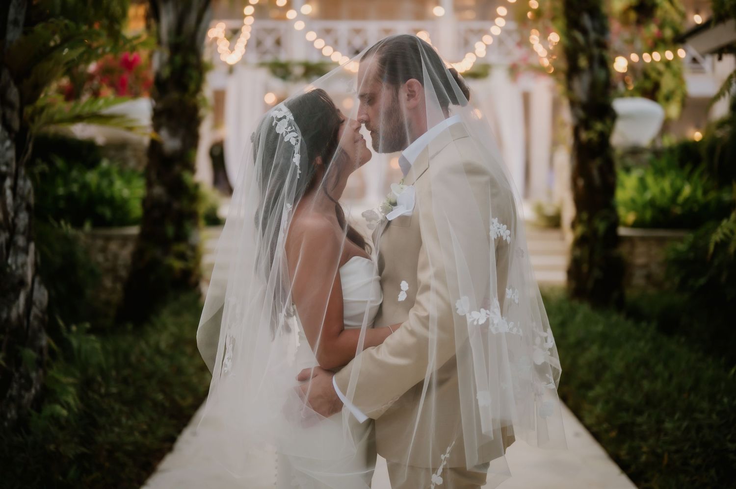 Newlyweds sharing a romantic kiss under the bride's veil in a garden with string lights.