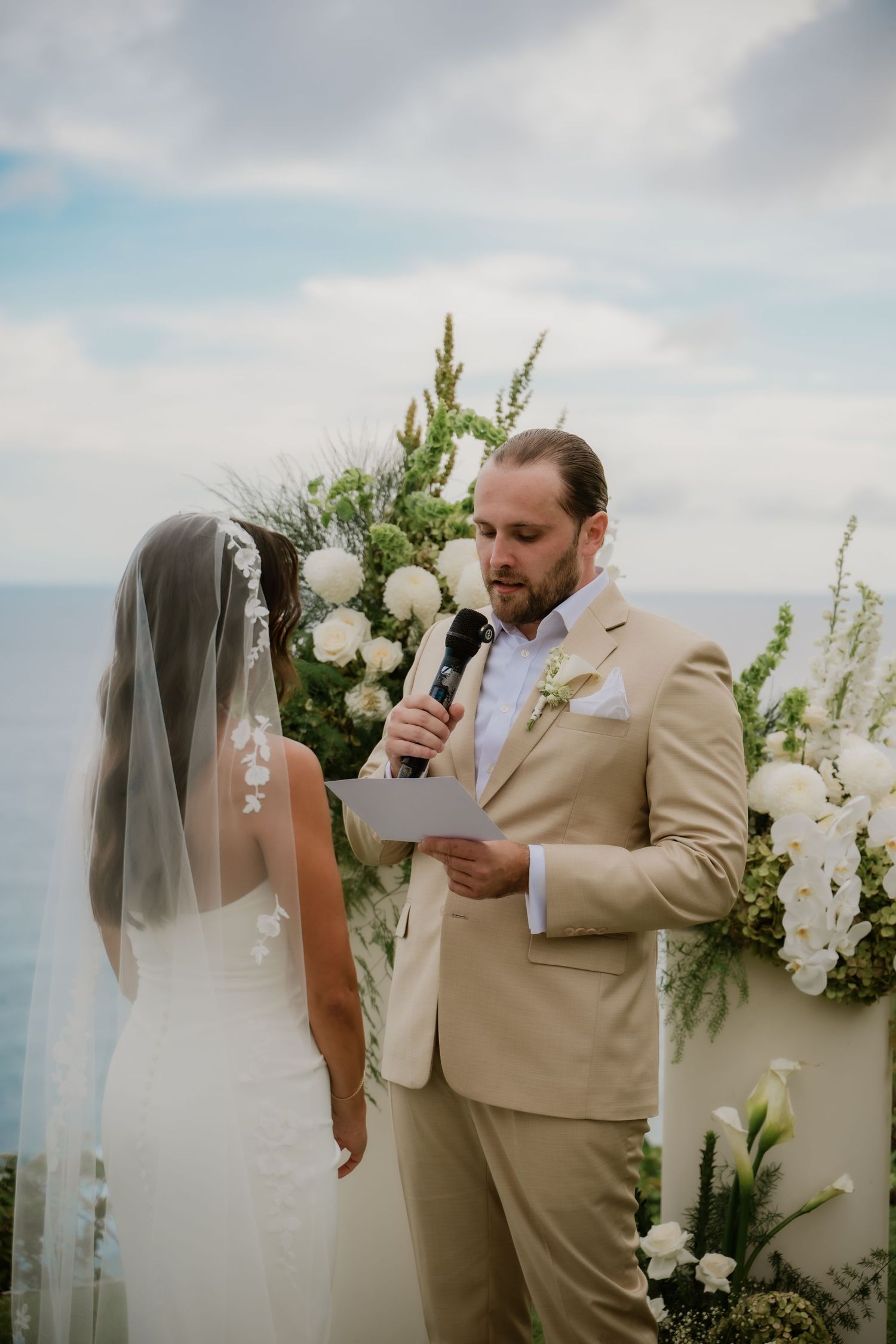 Groom in tan suit reading vows at outdoor ocean-view ceremony with white floral arch backdrop.