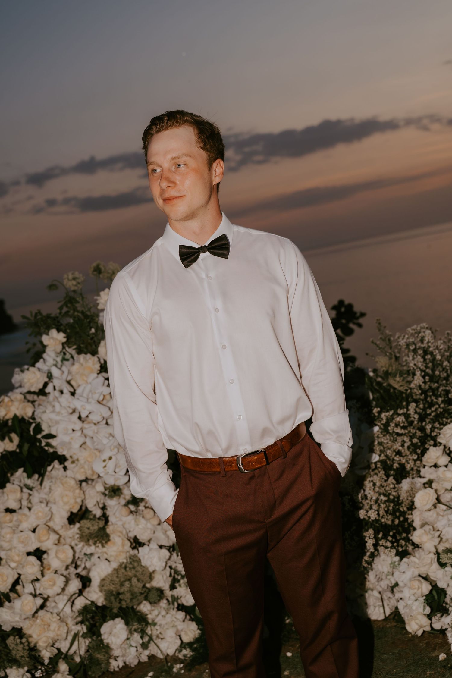 Groom in white shirt and black bow tie stands confidently among white flowers at sunset ceremony.