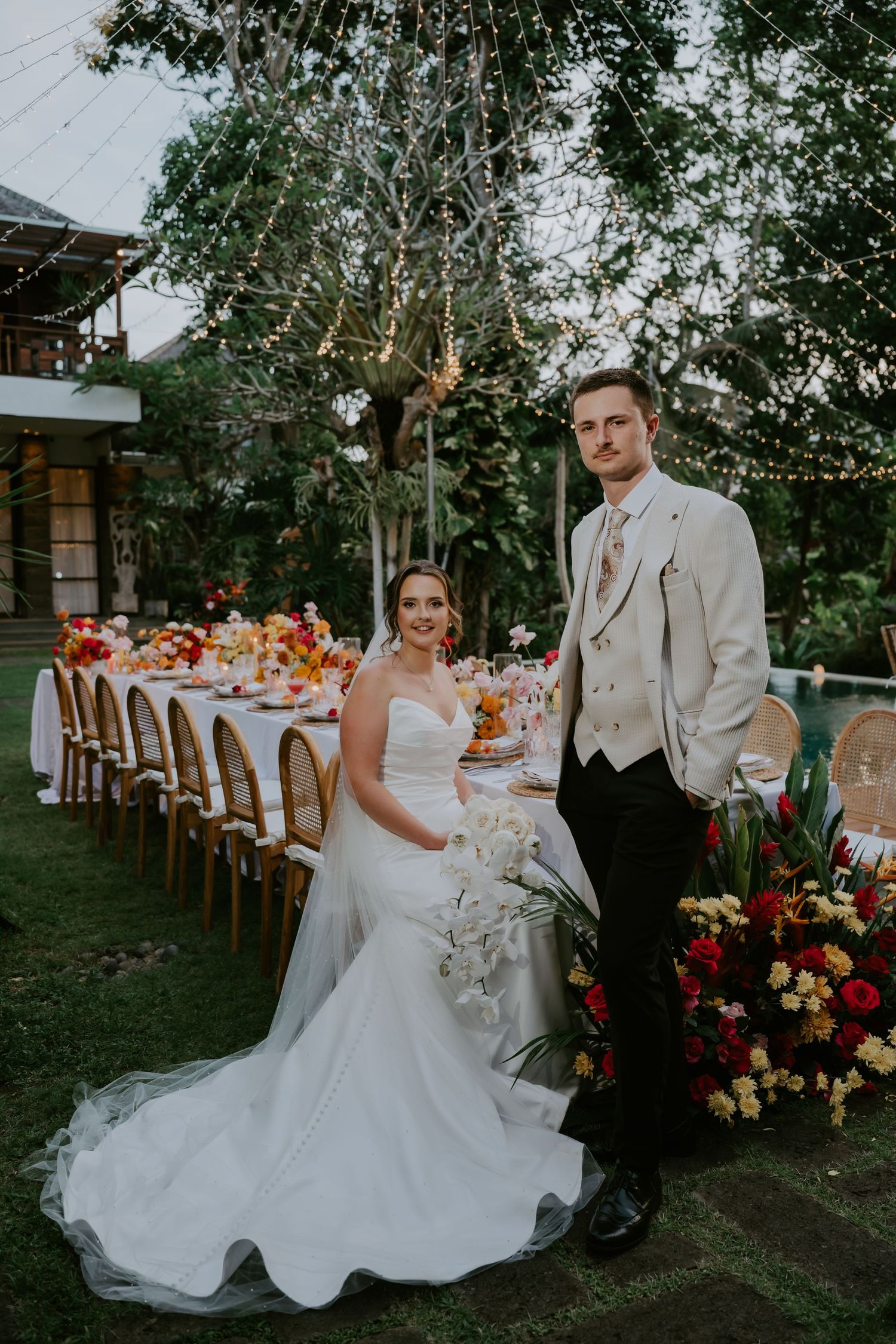 Bride and groom pose beside a long floral reception table with string lights and lush greenery at an outdoor wedding.