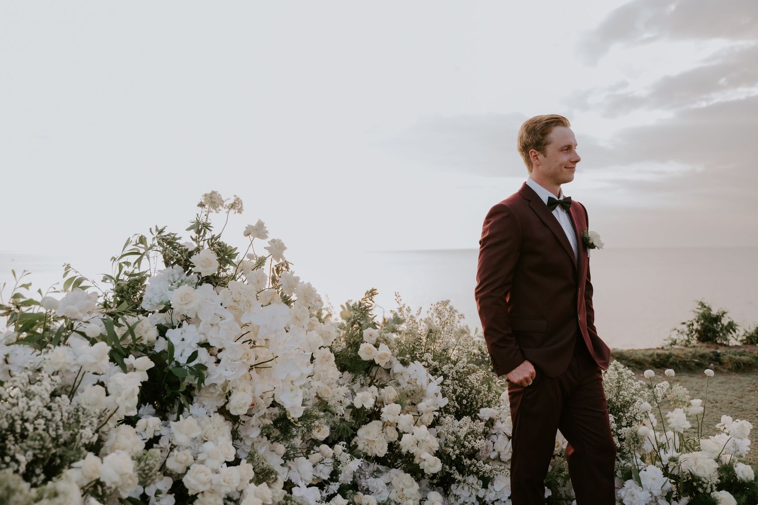 Groom in burgundy suit stands beside white floral arrangement with ocean backdrop at sunset wedding.