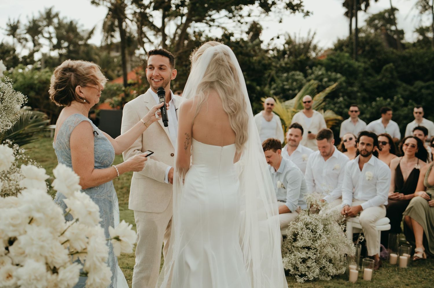 Groom in beige suit reads vows to bride in white gown before seated wedding guests outdoors.