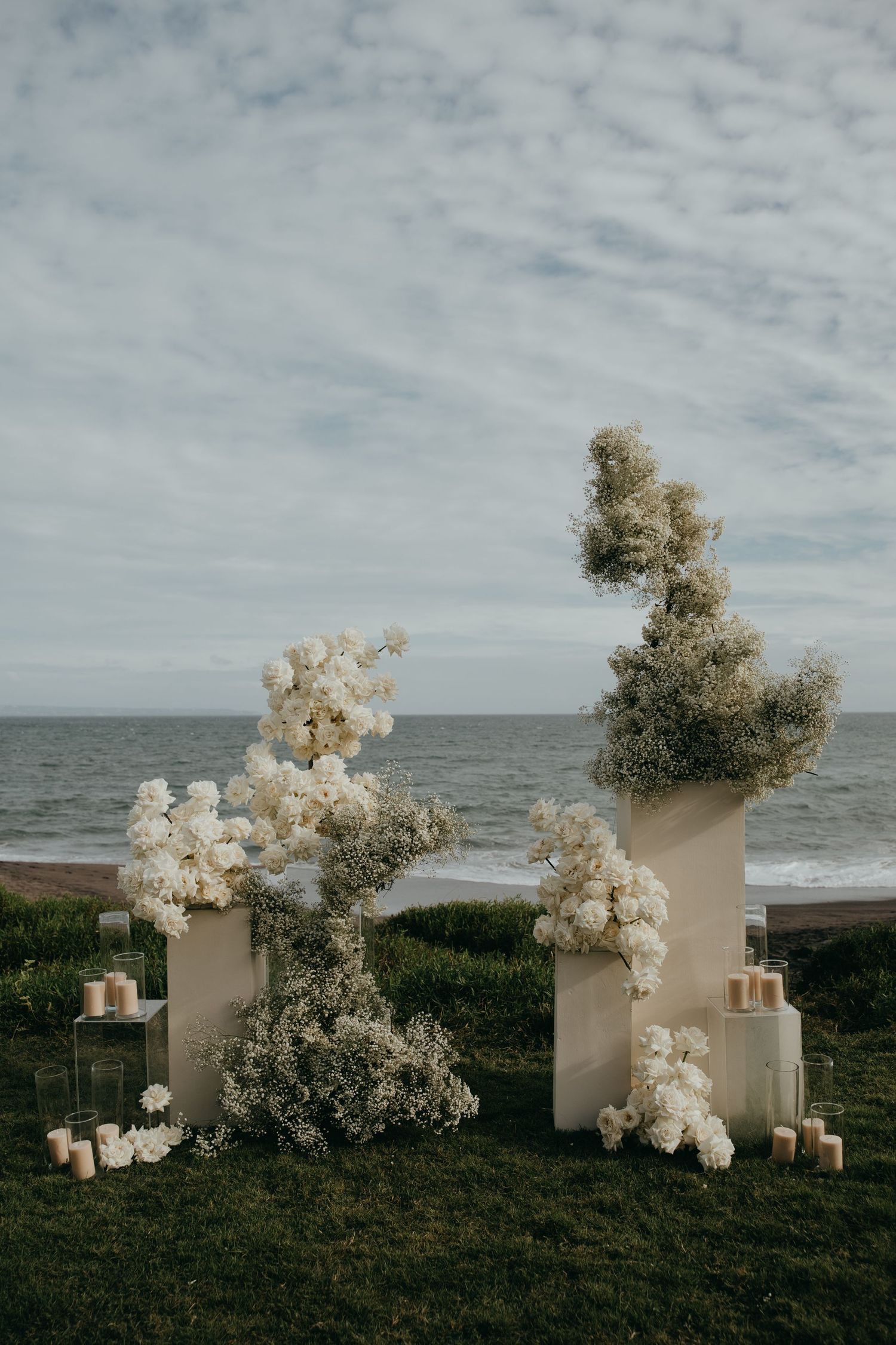 Two white floral pedestals with baby's breath and roses stand on grass overlooking the ocean at a coastal wedding ceremony.