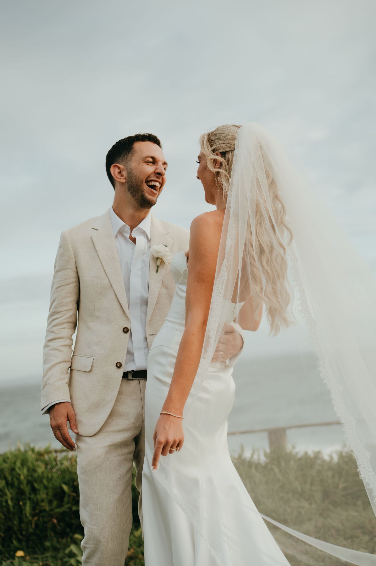 Laughing bride and groom in beige suit share a joyful moment outdoors on their wedding day.