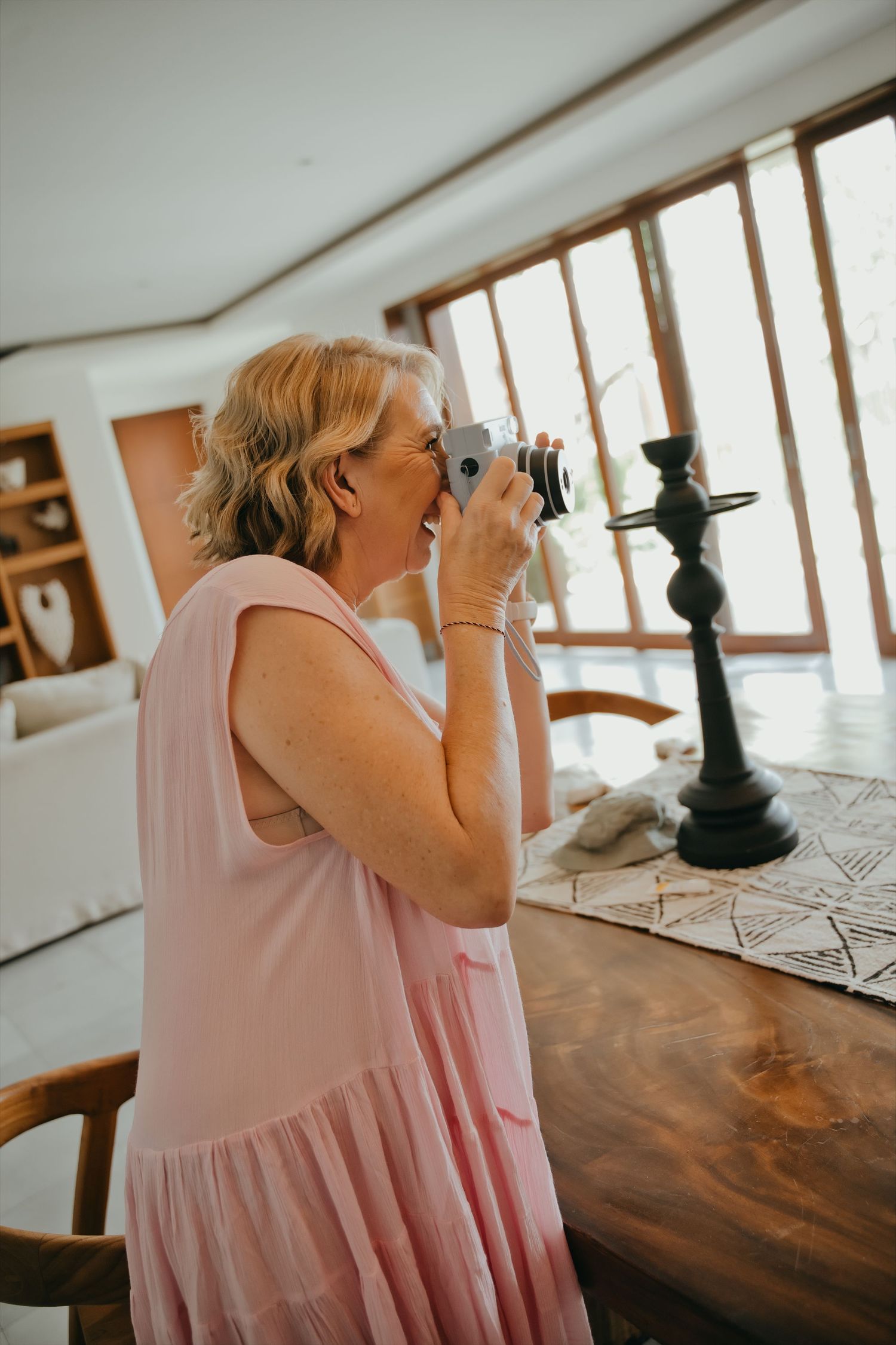 Woman in pink dress photographing with vintage camera indoors near wooden table and large windows.