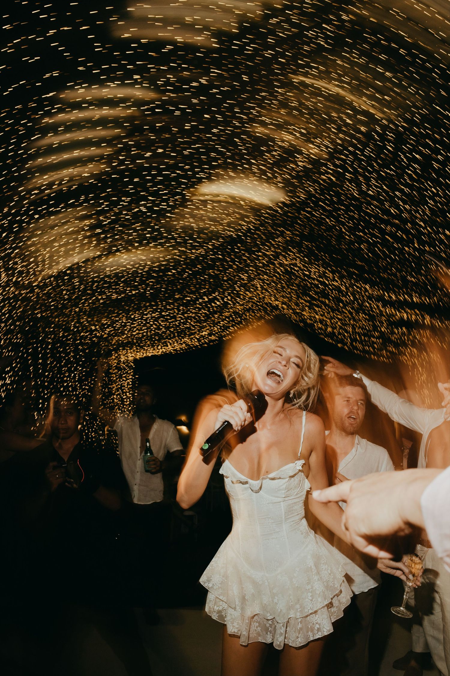 Bride singing into microphone in white mini dress under swirling fairy lights at wedding reception.