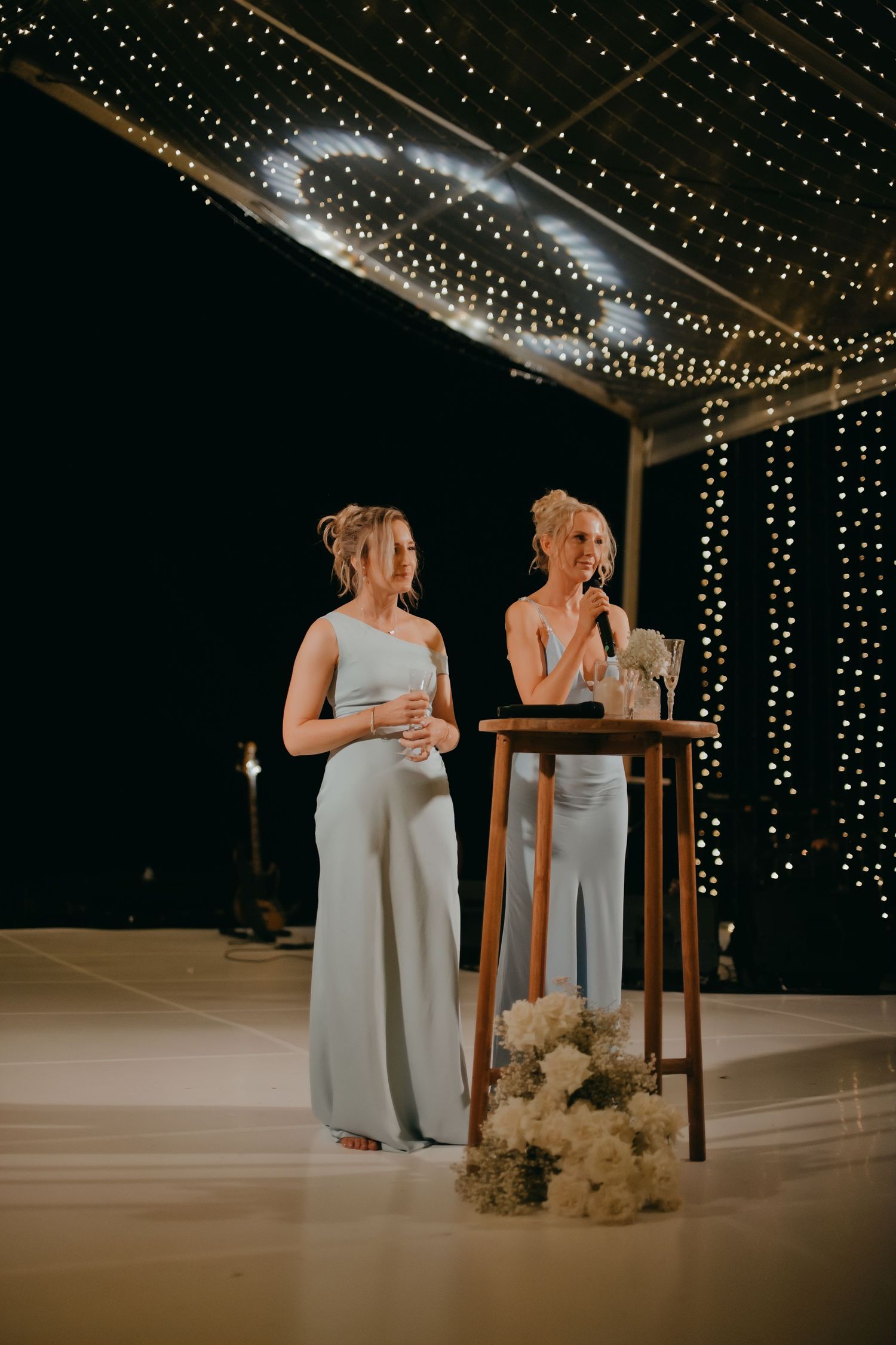Two bridesmaids in blue gowns delivering a speech at a fairy light wedding reception.