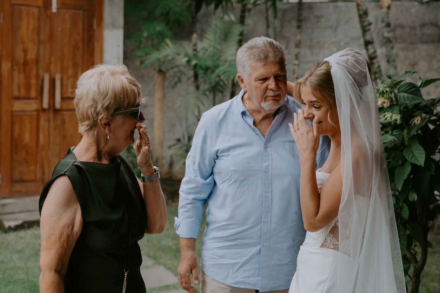 Emotional bride in wedding veil shares tearful moment with parents outdoors before wedding ceremony.