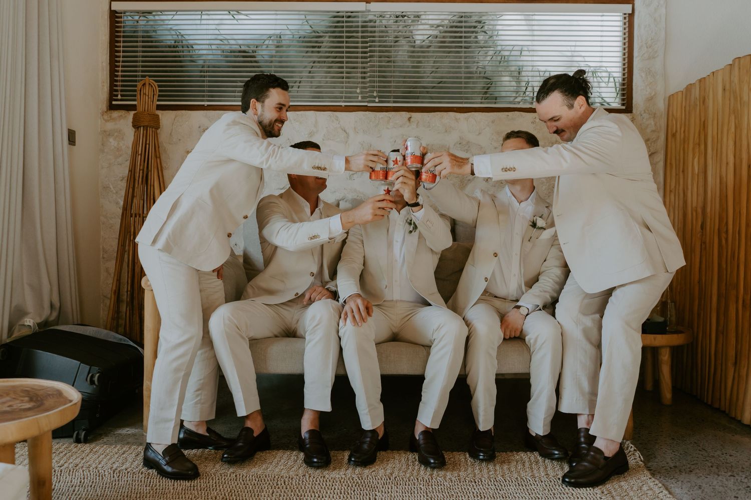 Four groomsmen in ivory suits toasting drinks together on a couch before the wedding.