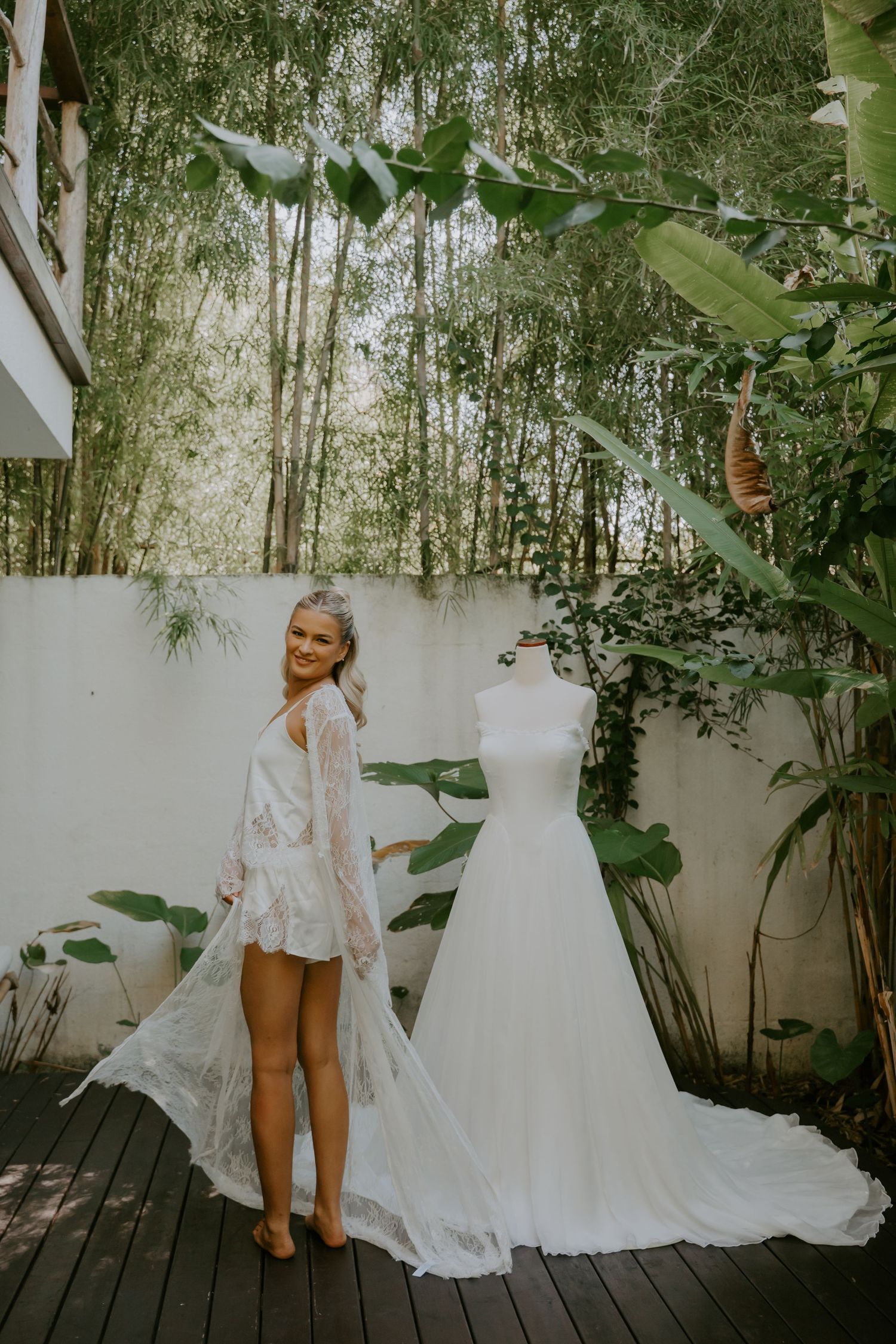 Bride in white lace robe stands beside her wedding dress on a mannequin in a tropical outdoor setting.