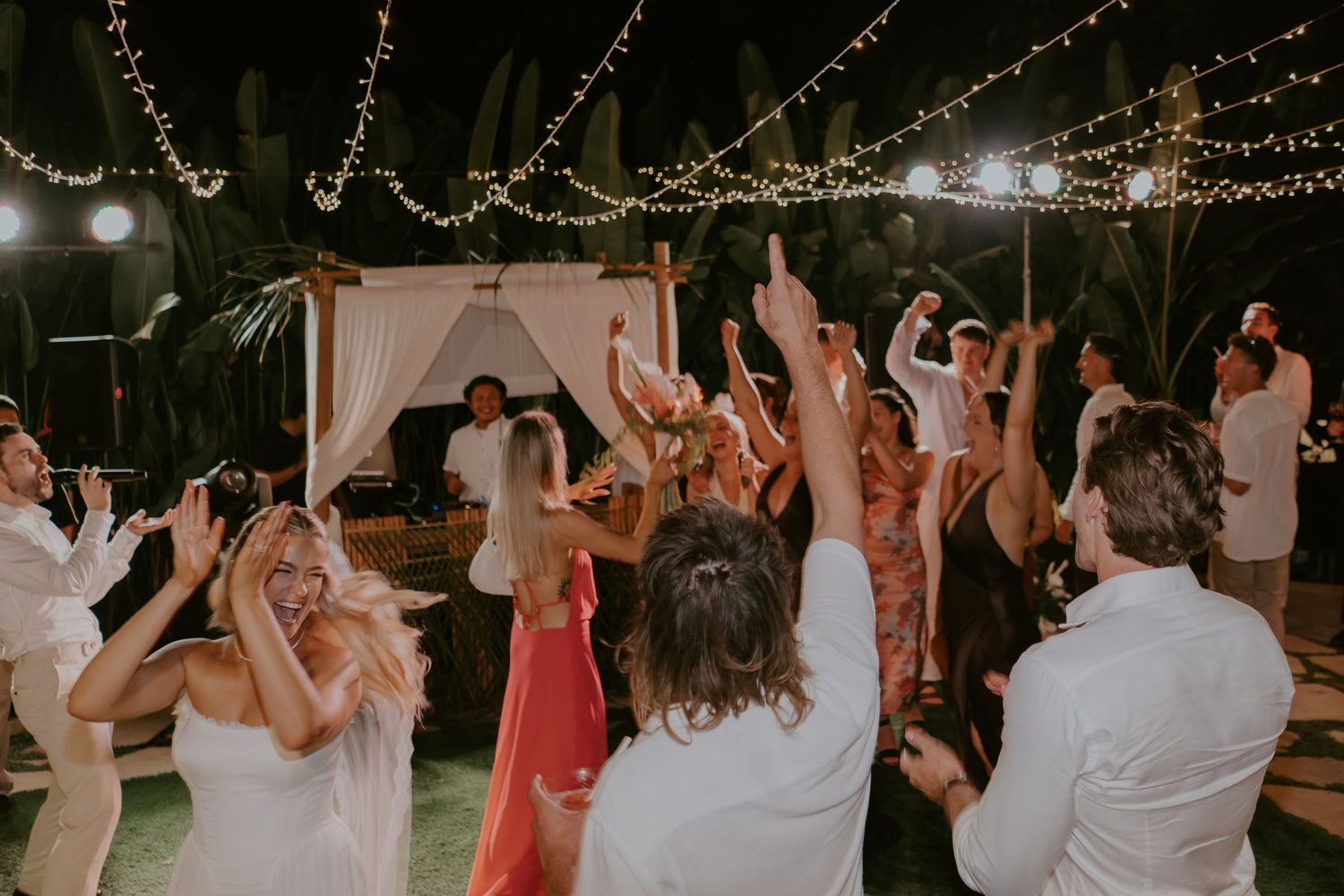 Wedding guests dancing with hands raised under string lights at an outdoor evening reception.