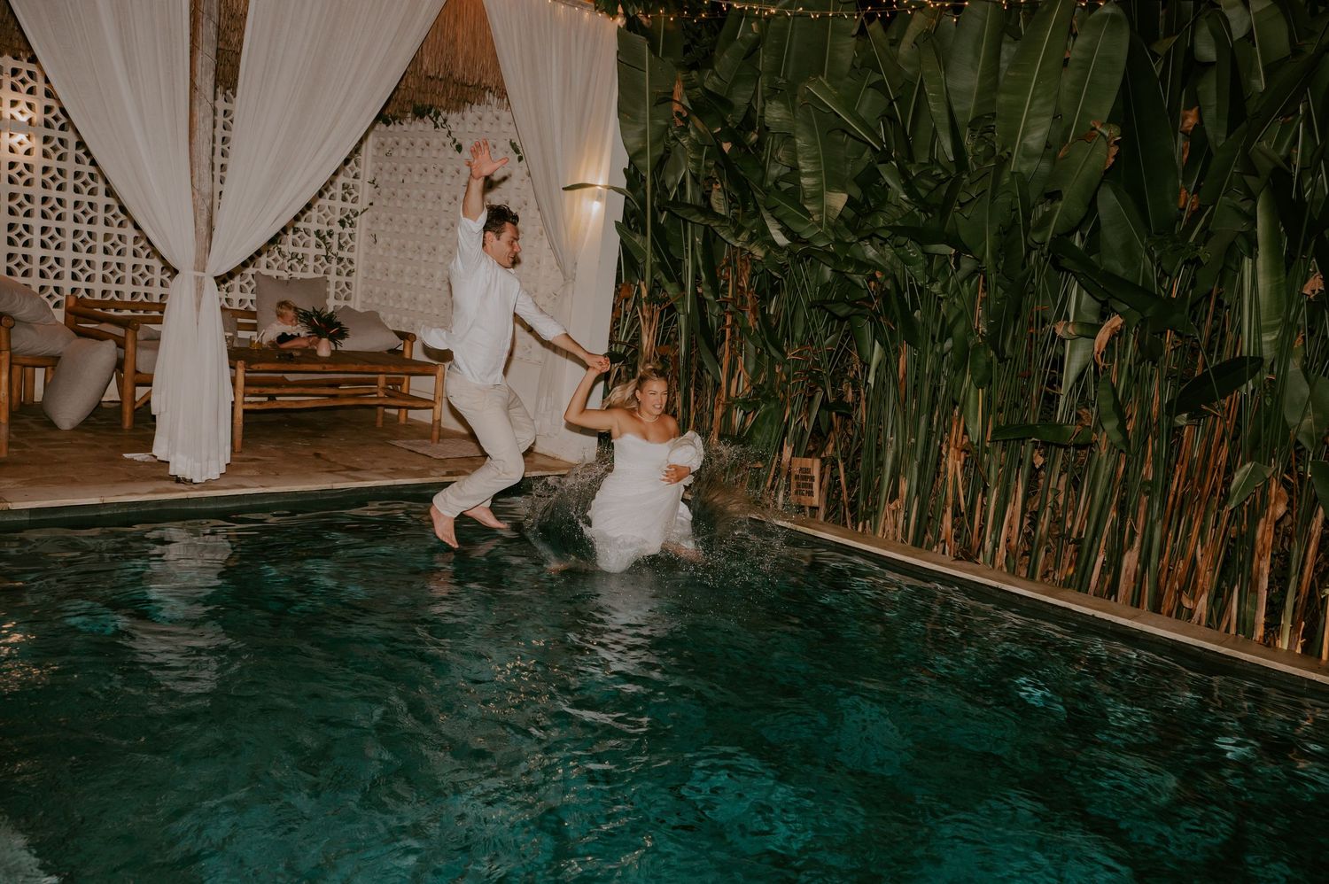 Bride and groom jumping into a pool together at night in their wedding attire.