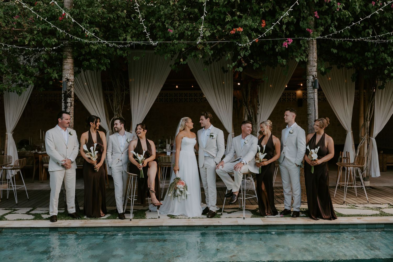 Wedding party poses poolside under string lights and lush greenery at tropical venue.